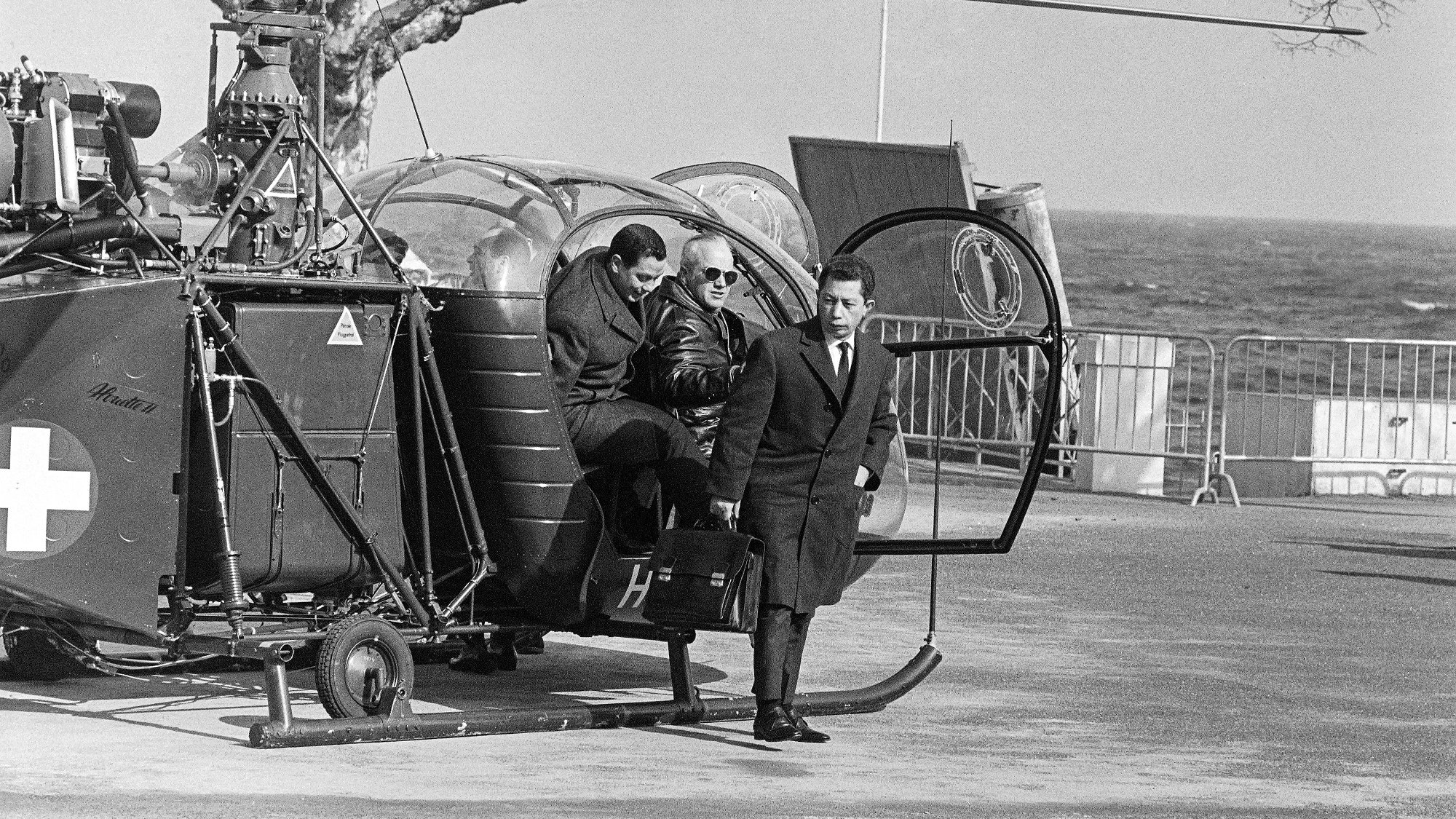 L'image montre une scène en noir et blanc avec un hélicoptère garé près de la mer. Deux personnes descendent de l'hélicoptère, tandis qu'une troisième personne les attend. On peut les voir porter des vêtements formels, ce qui suggère une certaine occasion. L'environnement est dégagé, avec une barrière visible en arrière-plan et des arbres dénudés. La mer se trouve en arrière-plan, ajoutant une touche de paysage côtier à la scène.