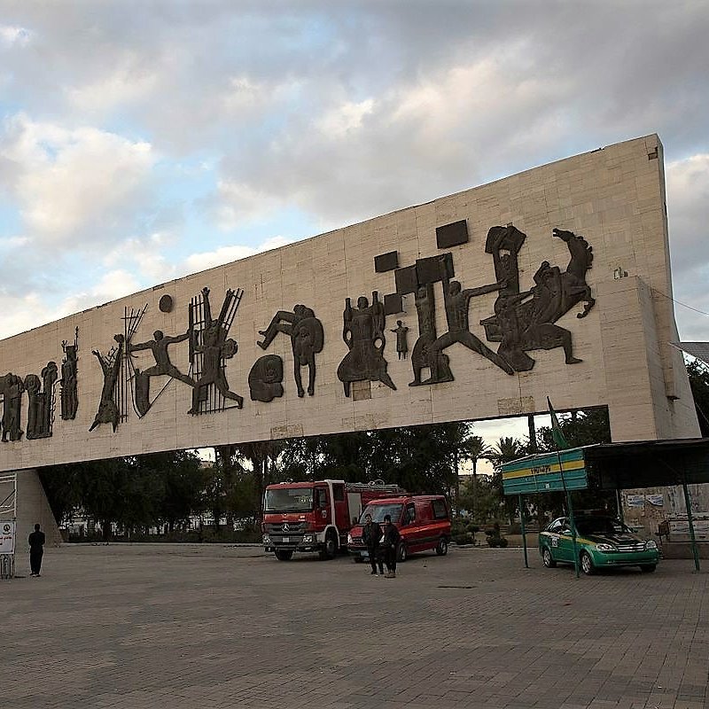 L'image montre une grande structure en béton surmontée de silhouettes découpées dans le matériau, représentant des figures humaines et des scènes de la vie quotidienne. Cette œuvre d'art moderne semble se dresser dans un espace public, entourée de véhicules et de personnes. Le ciel est nuageux, suggérant une ambiance calme et une lumière douce. En arrière-plan, on peut voir des bâtiments et des éléments d'urbanisme typiques d'une ville. L'ensemble évoque un mélange de culture et d'art, tout en étant intégré dans la vie citadine.