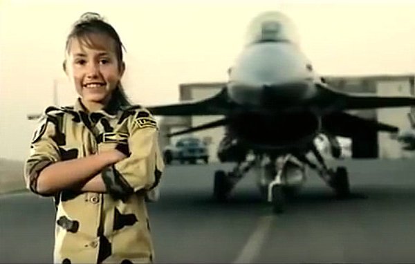 L'image montre une jeune fille souriante vêtue d'une tenue militaire camouflage. Elle se tient debout, les bras croisés, devant un avion de chasse. L'arrière-plan est flou, mettant l'accent sur l'avion, qui semble être un F-16. La scène dégage une ambiance de fierté et de bravoure, avec un ciel clair à l'horizon.