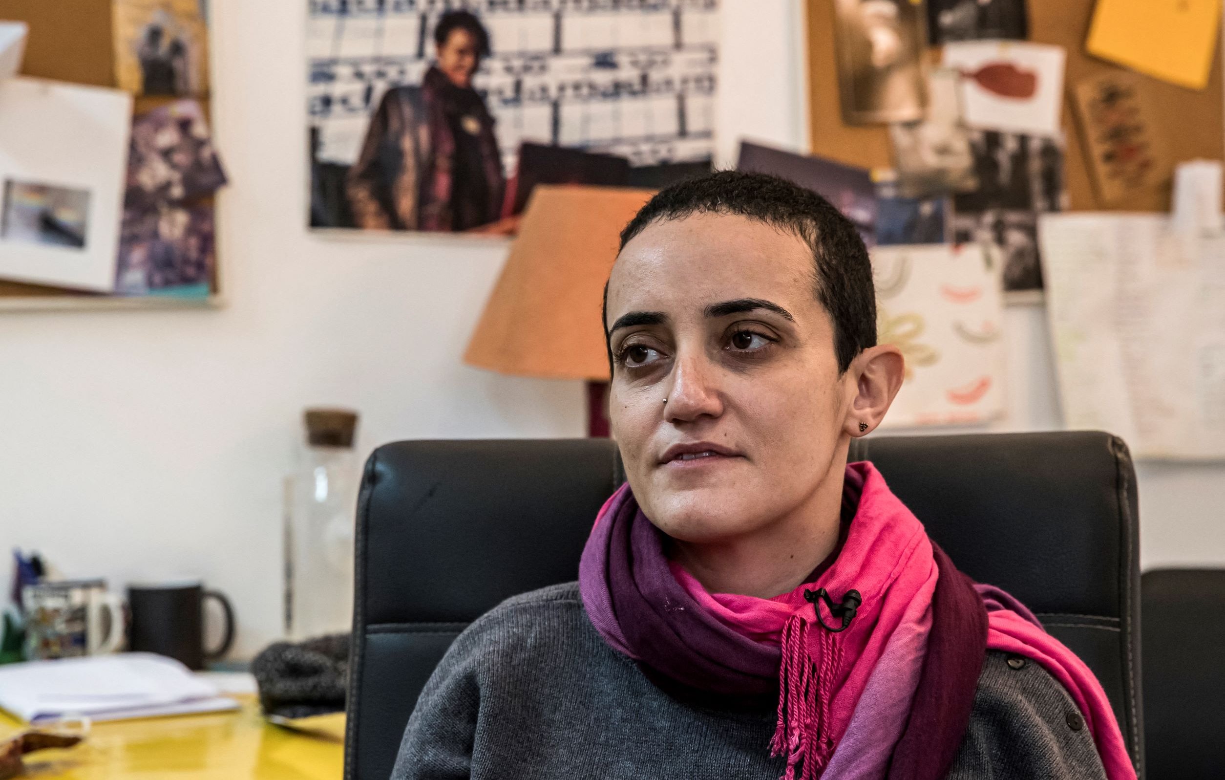 Je ne sais pas qui est présent sur cette image. Cependant, je peux te dire que la personne semble assise dans un bureau, portant un pull gris et une écharpe rose. On peut voir un environnement de travail avec des documents et des photos affichées en arrière-plan. L'éclairage donne une ambiance chaleureuse à la scène.