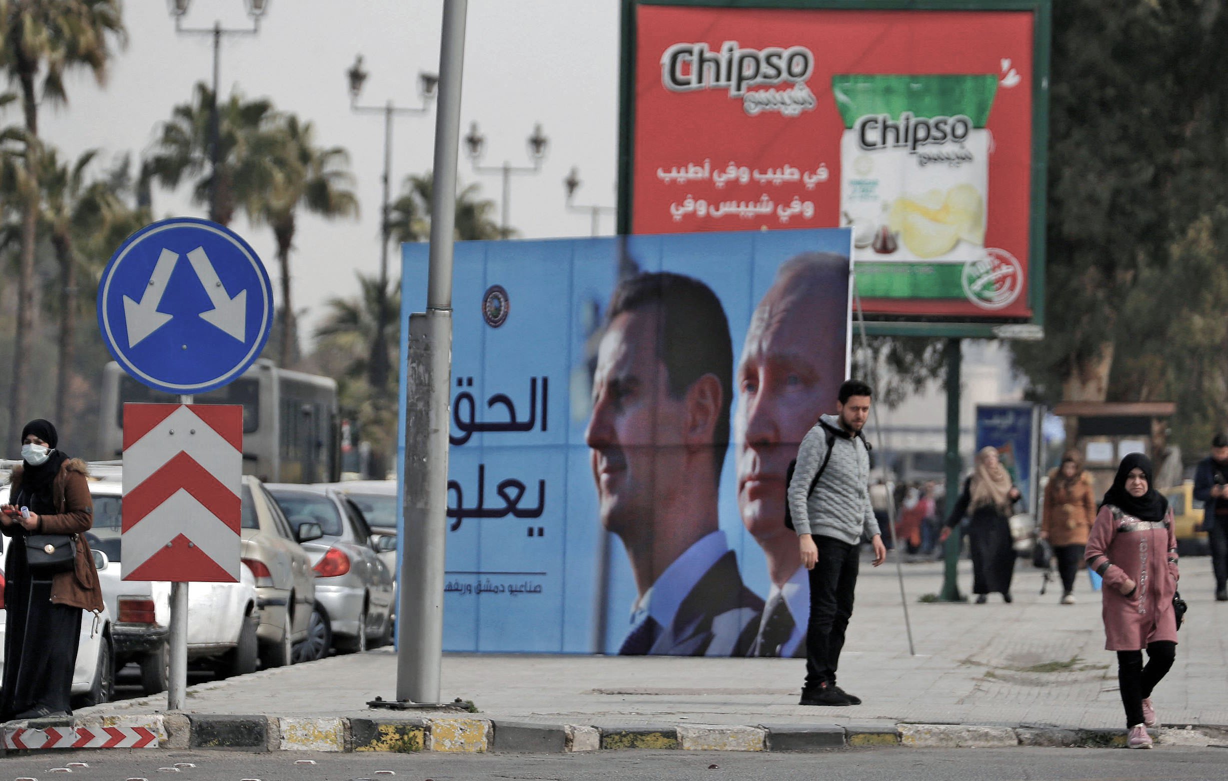 L'image montre une scène urbaine avec des panneaux de publicité. Il y a un panneau affichant des portraits de deux hommes, probablement des dirigeants, avec un message en arabe. À gauche, plusieurs personnes marchent le long de la route, tandis qu'à droite, des voitures sont garées. On peut également voir un rond-point typique et des palmiers en arrière-plan, ce qui donne un aspect typique d'une ville dans une région chaude. La publicité pour une marque de chips est visible au-dessus.