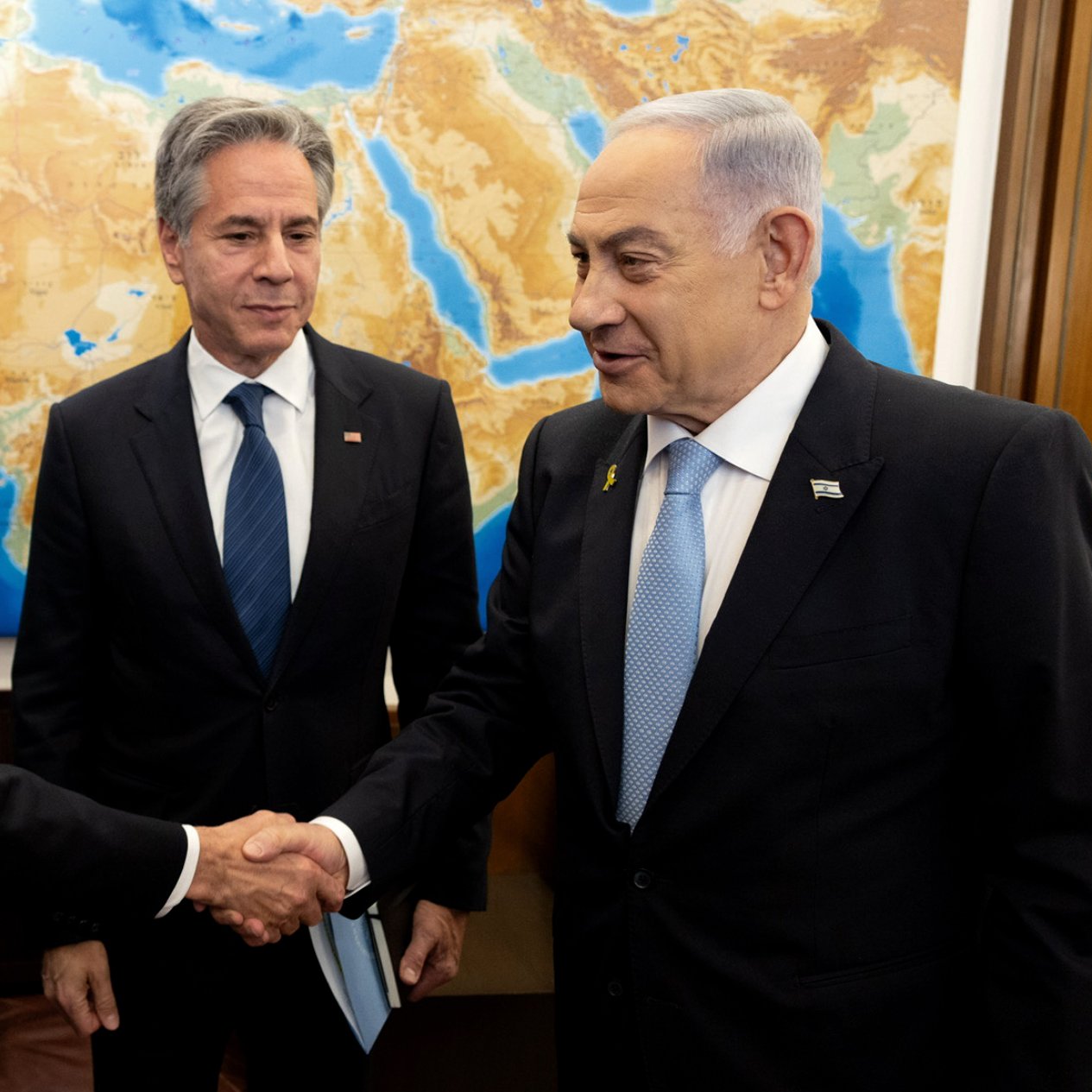 در تصویر، چند نفر در حال گپ‌وگفت هستند. دو نفر با هم دست می‌دهند و یکی دیگر در پس‌زمینه ایستاده است. پس‌زمینه شامل نقشه‌ای از منطقه خاورمیانه است. افراد در حال گفتگو به نظر می‌رسند و حالت چهره آن‌ها نشان‌دهنده تعامل مثبت است. لباس‌های رسمی آنها به اهمیت جلسه اشاره دارد.