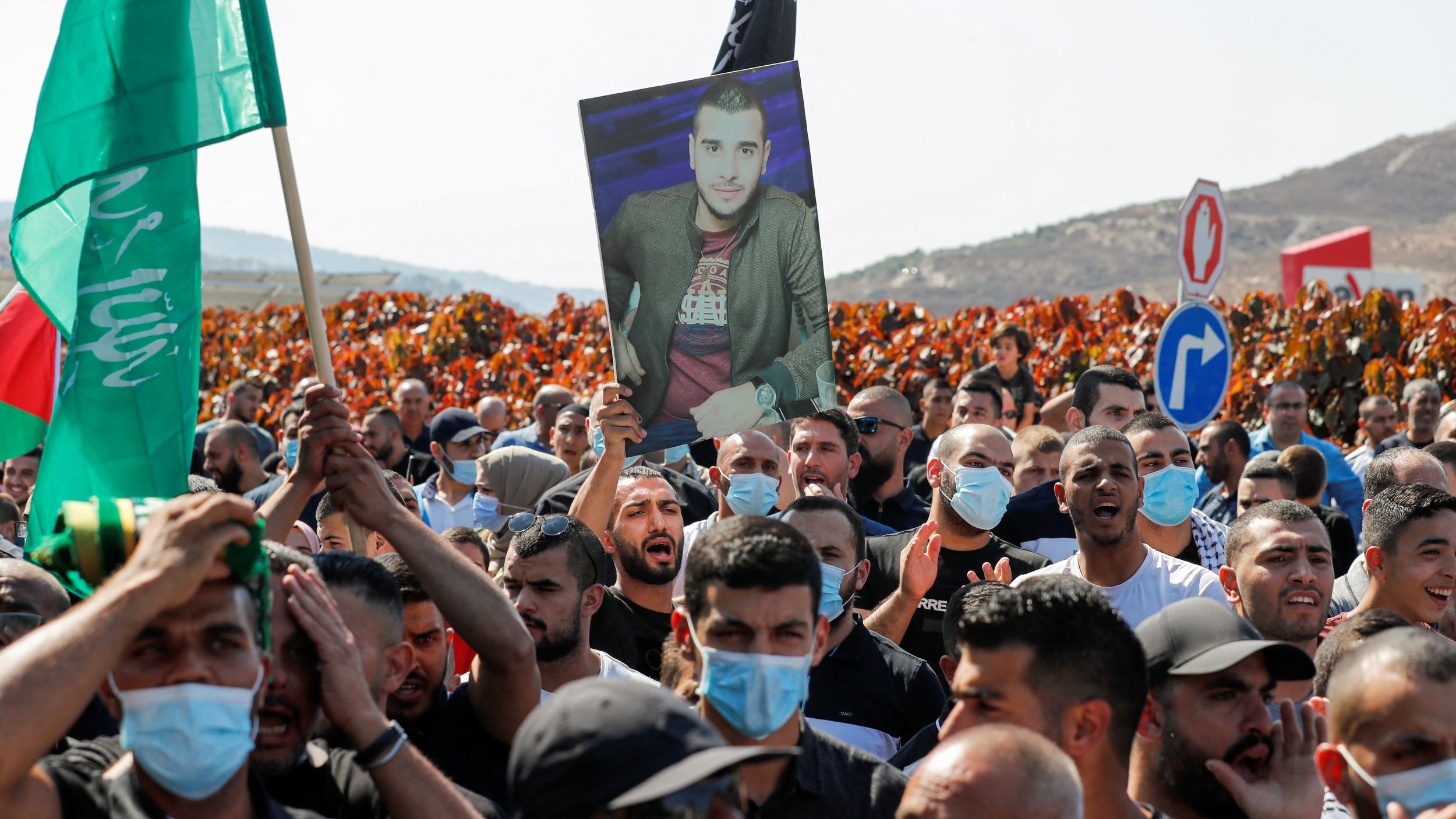 L'image montre une manifestation où une foule dense est présente. Au premier plan, des manifestants tiennent des drapeaux, dont un vert, et certains agitent des pancartes. Parmi eux, une personne porte un grand portrait d'un individu, visiblement en hommage ou en soutien. Les manifestants semblent engagés et déterminés, et ils portent tous des masques sanitaires, indiquant un contexte de pandémie. En arrière-plan, des collines se dessinent, avec des panneaux de signalisation visibles. L'atmosphère semble chargée d'émotion et de revendications.