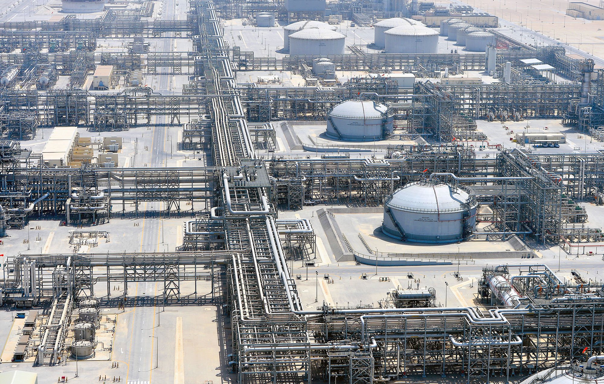 L'image montre une vue aérienne d'une installation industrielle, vraisemblablement une raffinerie ou un complexe pétrolier. On peut y voir des réservoirs de stockage de forme sphérique, entourés de nombreux tuyaux et structures métalliques. L'environnement est apparemment désertique, avec des terrains arides et peu de végétation. Plusieurs installations industrielles sont visibles, indiquant une activité de traitement ou de transport de matières pétrolières.