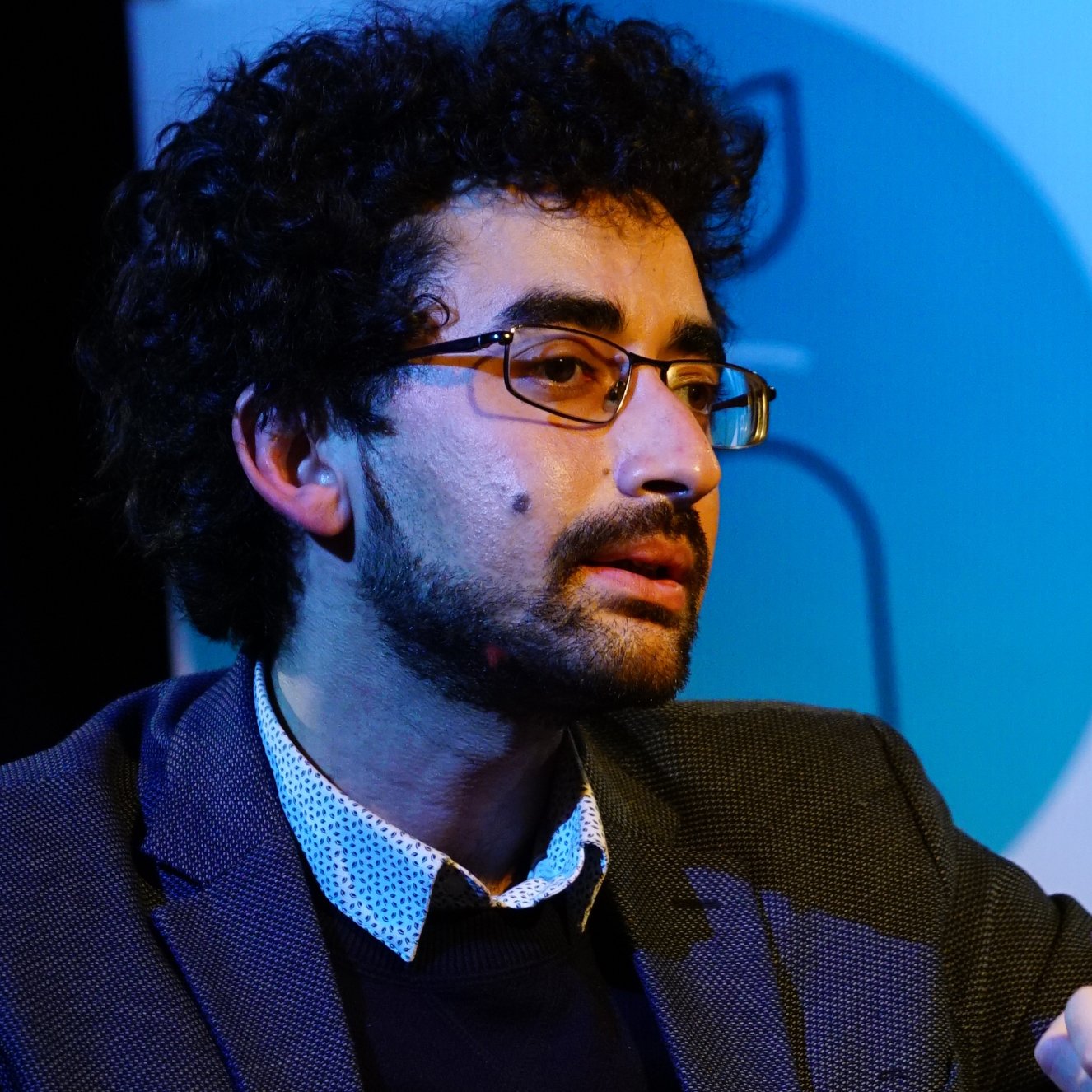 Homme avec des lunettes, cheveux bouclés, en discussion, vêtu d'une veste.