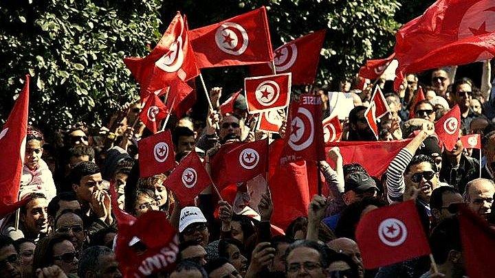L'image montre une foule de personnes rassemblées, agitant des drapeaux tunisiens rouges avec un cercle blanc et une étoile à cinq branches. Les manifestants semblent animés, levant les bras en signe de solidarité ou de protestation. On peut voir un paysage urbain en arrière-plan avec des arbres, ce qui suggère un rassemblement en plein air. L'atmosphère semble énergique et engagée, reflétant un moment de mobilisation collective.