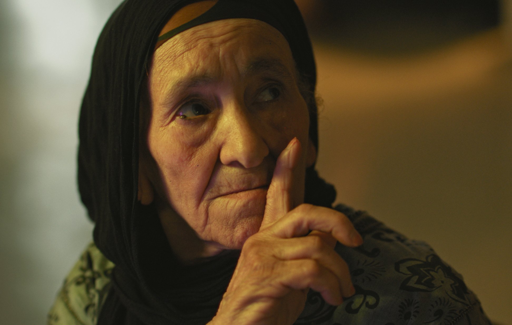 L'image montre une femme âgée avec un regard pensif. Elle porte un foulard noir qui encadre son visage. Son expression est sérieuse et elle semble faire un geste de silence avec son doigt posé sur ses lèvres. La lumière dans le fond est douce, créant une ambiance calme et introspective. Les détails de son visage reflètent une certaine sagesse et vécu.