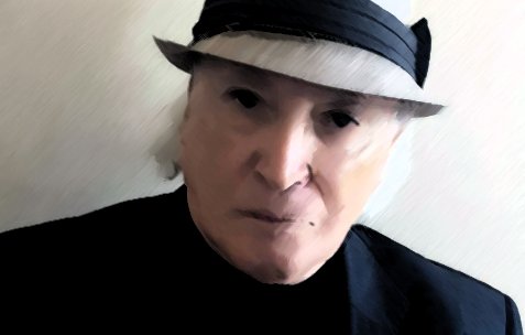 الصورة تظهر شخصًا يرتدي قبعة رمادية مع شريط داكن، وهو يرتدي سترة داكنة أيضًا. لديه ملامح وجه جادة، والخلفية تبدو بسيطة وغير مزدحمة، مما يركز الانتباه على تعبيره وملابسه. توزيع الإضاءة في الصورة يبرز ملامحه بشكل واضح.