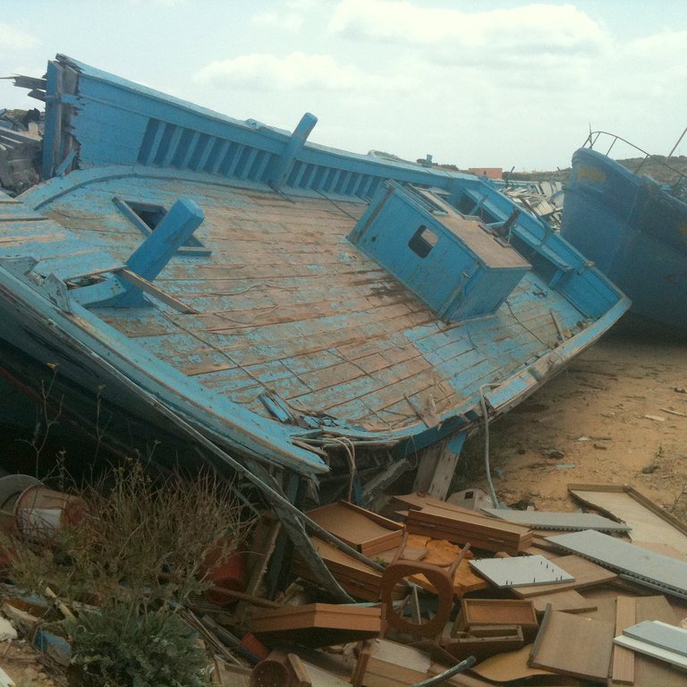 L'image montre des bateaux endommagés, apparemment abandonnés sur une terre aride. Les bateaux, de couleur bleu clair, sont inclinés sur le côté, avec des parties de leur structure exposées. On peut voir des débris et des objets éparpillés autour, ainsi qu'une végétation rudimentaire qui pousse dans le sol. Le ciel est nuageux, créant une atmosphère mélancolique. L'ensemble dégage une impression de désolation et d'abandon.