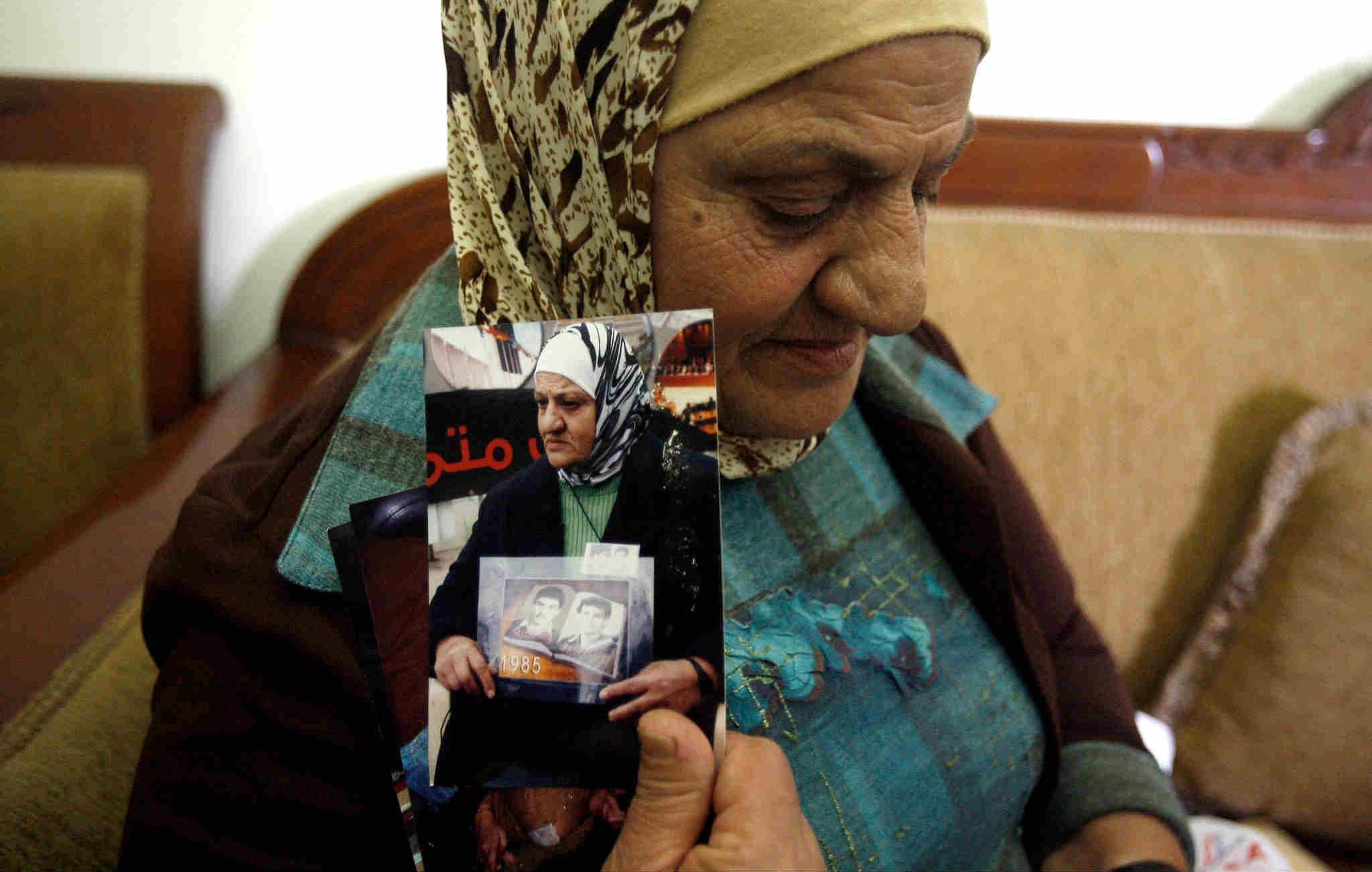 L'image montre une femme âgée portant un foulard, assise sur un canapé. Elle tient des photos en noir et blanc dans ses mains. Son expression semble empreinte de nostalgie ou de tristesse, indiquant peut-être qu'elle évoque des souvenirs douloureux. L'environnement semble simple, avec des murs clairs et un décor sobre en arrière-plan. La scène transmet une forte charge émotionnelle, suggérant un lien personnel avec les images qu'elle tient.