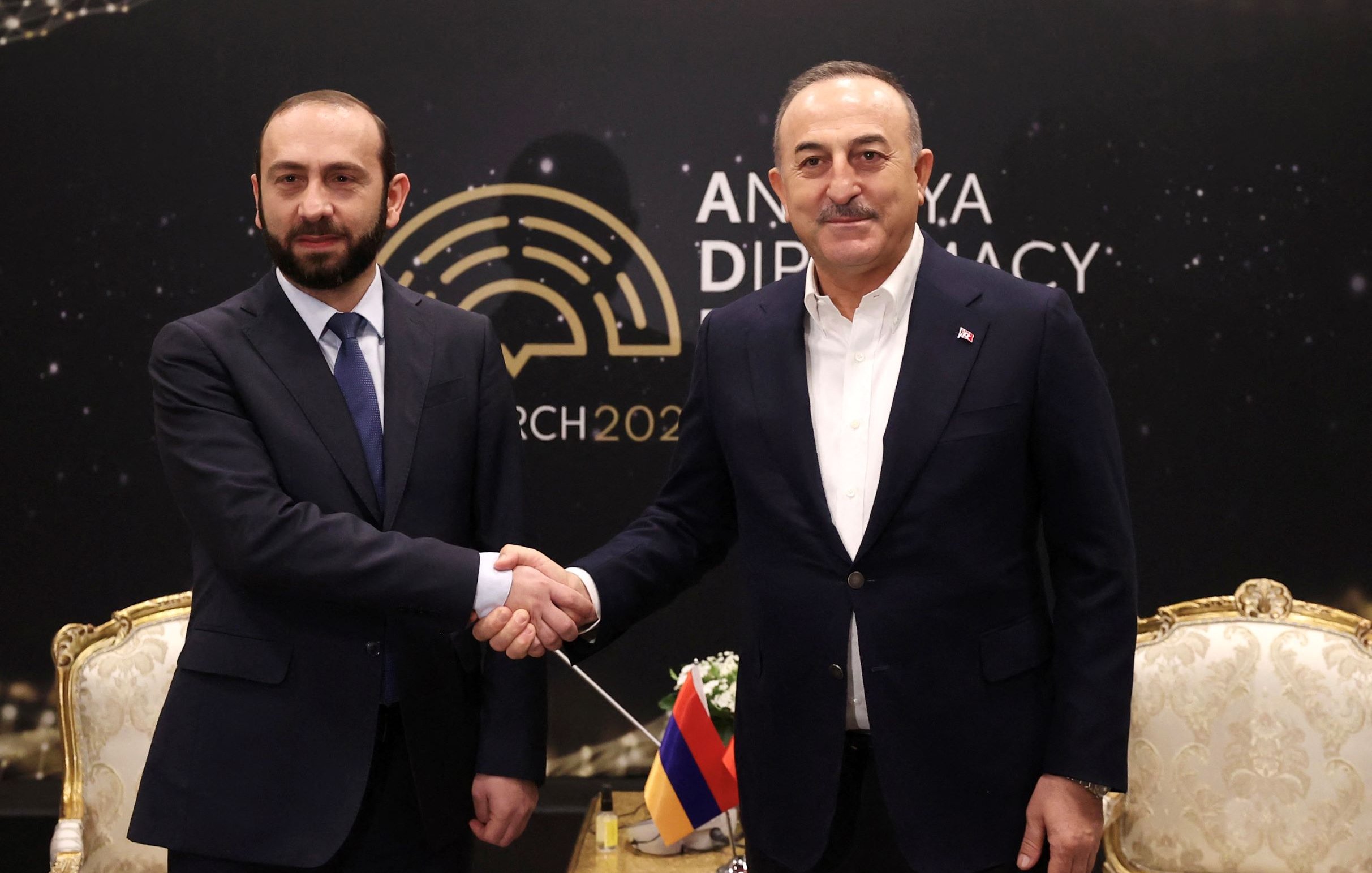 L'image montre deux hommes se serrant la main, probablement en signe de coopération ou de négociation. Ils sont vêtus de costumes formels. L'arrière-plan présente le logo d'un événement lié à la diplomatie, avec la mention "Ankara Diplomacy 2023". L'ambiance semble être celle d'une rencontre officielle.