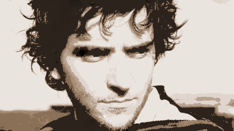 L'image présente un homme avec des cheveux bouclés et une expression sérieuse. Son visage est éclairé d'une manière qui met en valeur ses traits, et il semble regarder au loin. Le fond est flou, ce qui met l'accent sur son visage. La palette de couleurs est principalement en nuances sépia, ce qui donne une atmosphère nostalgique ou intemporelle à l'image.