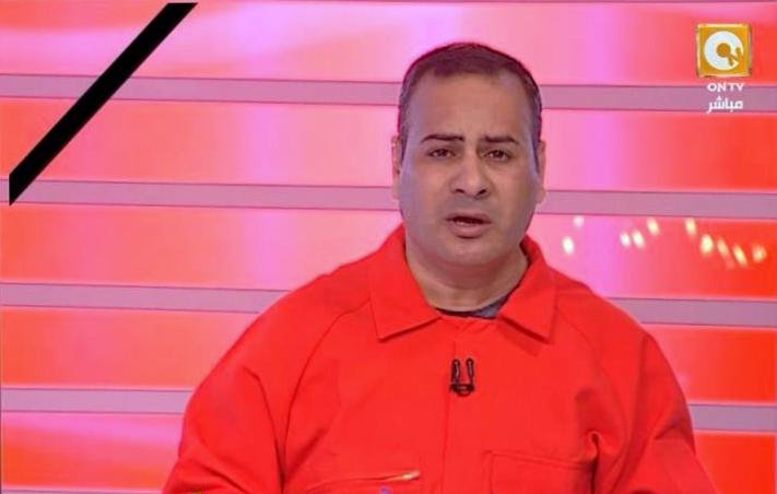 L'image montre un homme en train de parler devant un fond rouge avec des lignes lumineuses. Il porte une combinaison orange et semble être en train de faire une présentation ou de commenter quelque chose. Son expression faciale est sérieuse et il tient un stylo dans sa main. Le décor et son apparence suggèrent qu'il s'agit d'un animateur ou d'un commentateur à la télévision.