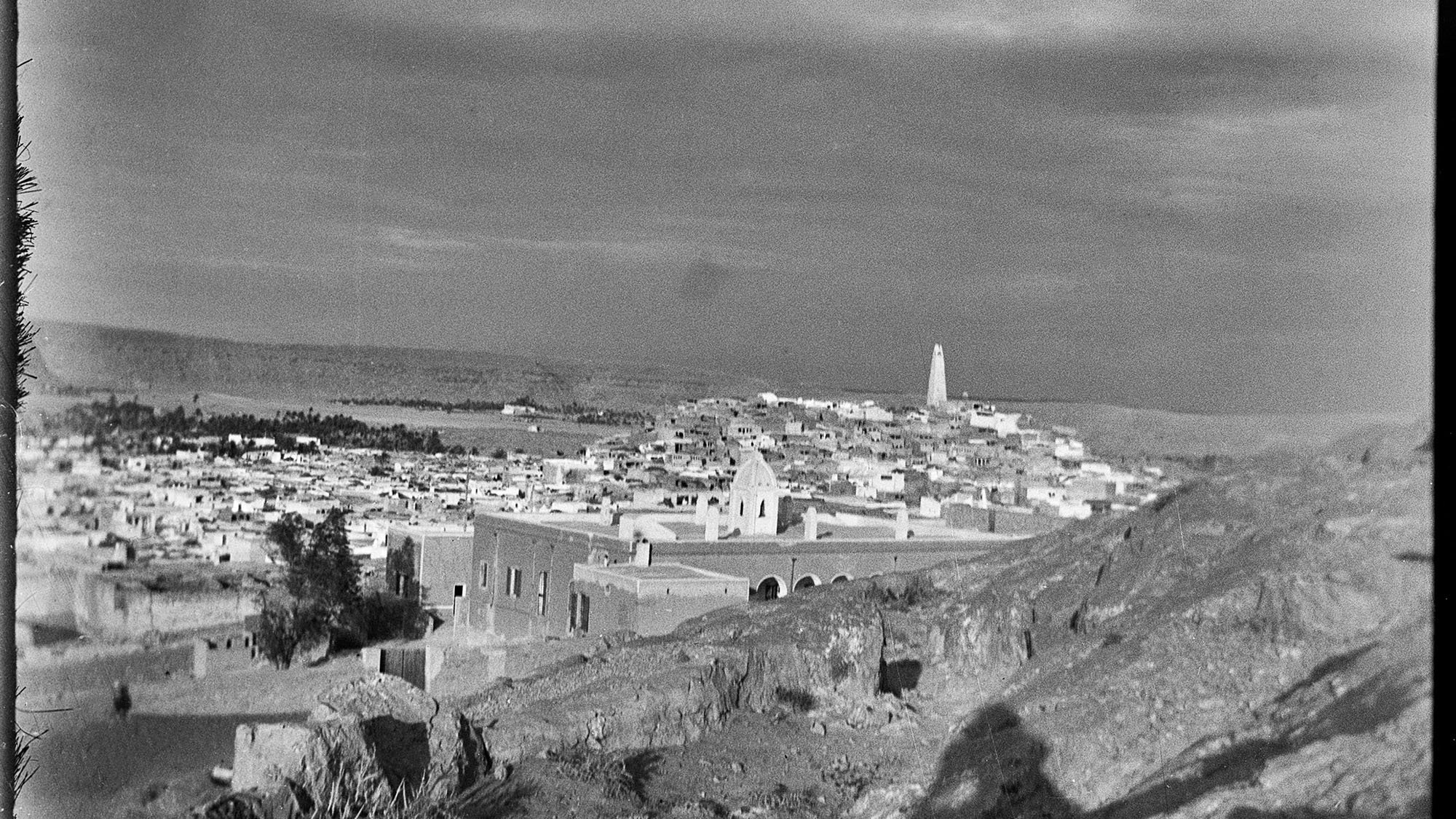 L'image montre un paysage urbain, probablement d'une ville historique. On peut voir des maisons blanches et des structures architecturales qui s'étendent à travers la vallée. À l'arrière-plan, un minaret s'élève, suggérant une présence religieuse. Le ciel semble nuageux, ce qui ajoute une ambiance dramatique à la scène. Le terrain est rocailleux au premier plan, et l'éclairage donne une impression de nostalgie, typique des photographies anciennes.