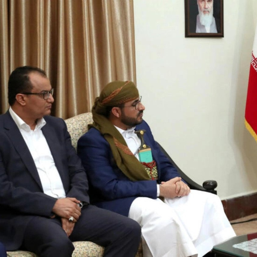 در این تصویر، دو مرد در حال گفتگو با یک شخصیت دیگر هستند. آن‌ها در یک مکان رسمی نشسته‌اند و تصاویر و پرچم‌هایی در پس‌زمینه دیده می‌شود. یکی از مردان لباس سنتی پوشیده و دیگری لباس رسمی دارد. شخصیت دیگر که صحبت می‌کند، عمامه و لباس سنتی ایران را به تن دارد. فضای گفتگو نشان‌دهنده یک جلسه ملاقات رسمی است.