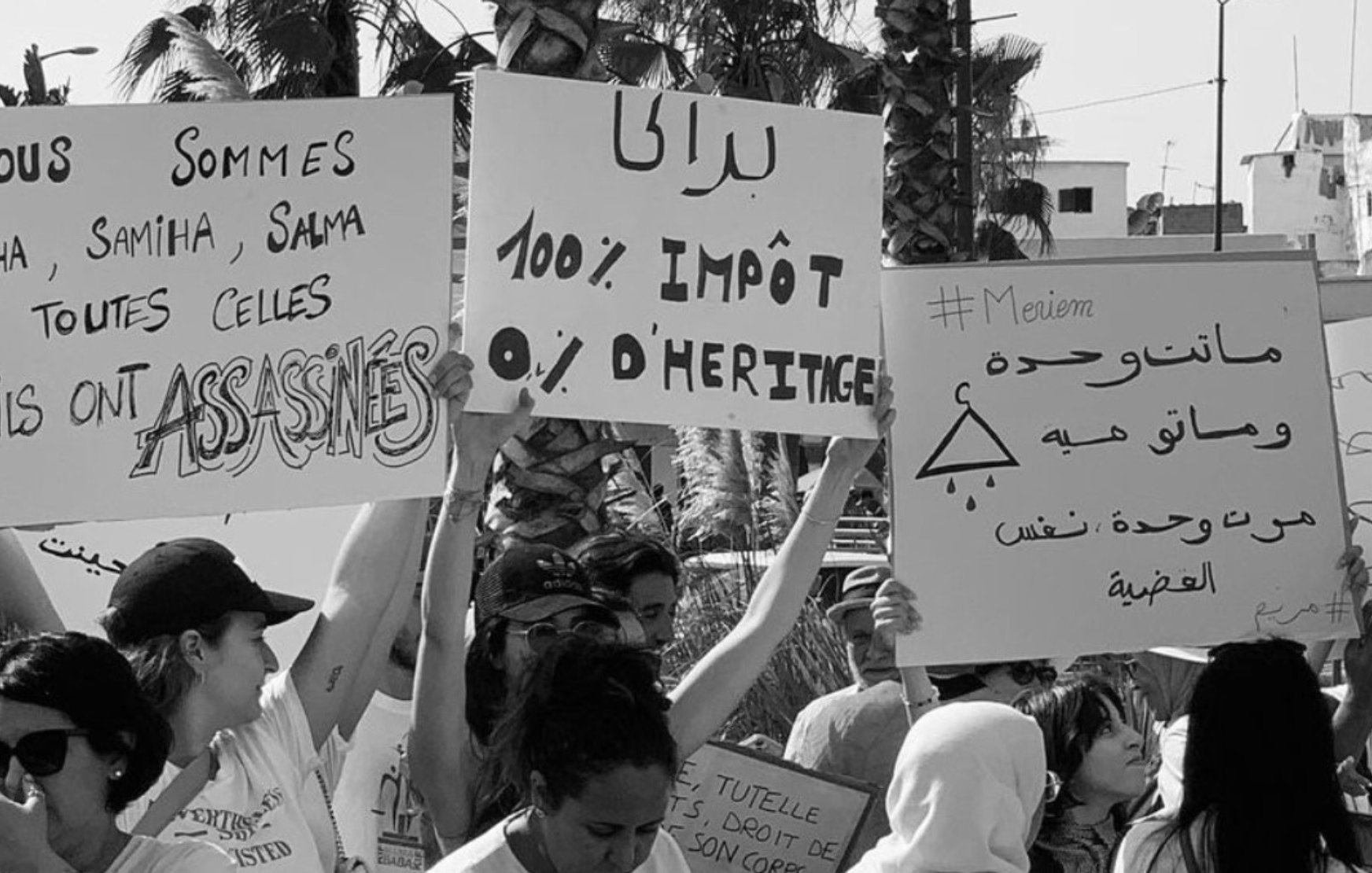 L'image est en noir et blanc et montre un rassemblement de personnes tenant des pancartes. Certaines des pancartes portent des messages en français comme : - "Nous sommes Aïcha, Samiha, Salma et toutes celles qu'ils ont ASSASSINÉES" - "100% IMPÔT 0% D'HÉRITAGE" Une autre pancarte comporte des inscriptions en arabe et le hashtag "#Meviem." Les personnes dans l'image semblent être engagées dans une manifestation ou une protestation concernant les droits des femmes et les questions de violence ou d'injustice. On peut également voir quelques palmiers et des bâtiments en arrière-plan, ce qui suggère que la scène se déroule en plein air dans un lieu urbain.