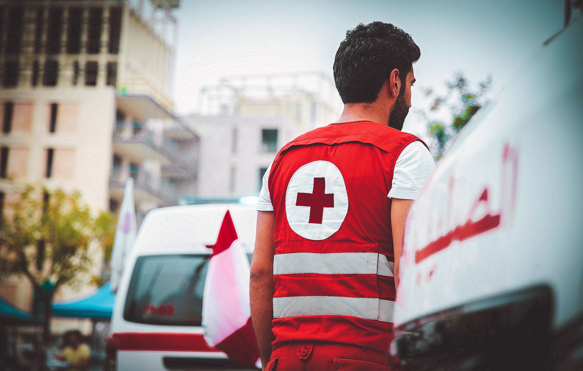 L'image montre un homme portant une veste rouge avec une croix blanche, typique des membres d'une organisation de secours, comme la Croix-Rouge. Il se tient de dos, observant une scène à proximité de véhicules de secours. En arrière-plan, on aperçoit des bâtiments modernes et des drapeaux, ce qui pourrait indiquer un environnement urbain. L'atmosphère semble calme, mais le contexte suggère une implication dans des missions sanitaires ou d'assistance.