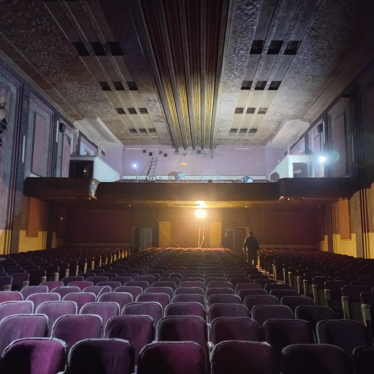 L'image montre l'intérieur d'un théâtre ancien. On peut voir des rangées de sièges en velours, qui semblent un peu usés, dirigées vers une scène surélevée. La scène est éclairée par une lumière vive, tandis que le reste de la salle est dans une ambiance plus sombre. On aperçoit également des détails architecturaux au plafond et sur les murs, qui évoquent un style vintage. Un homme se tient debout au fond, ajoutant une touche humaine à cet espace, qui dégage une atmosphère à la fois nostalgique et mystérieuse.