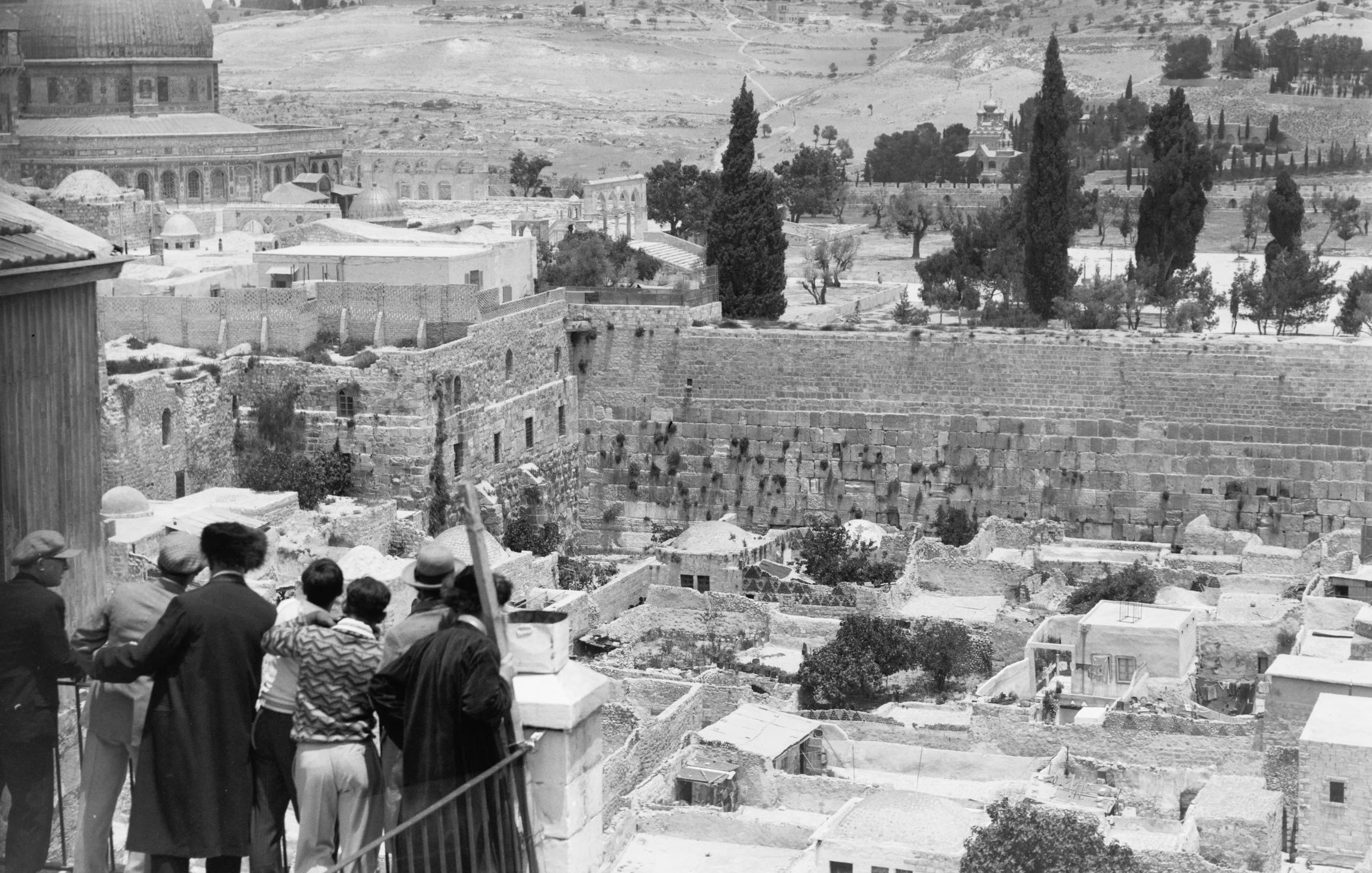 L'image montre une vue panoramique d'un paysage urbain historique. Au premier plan, un groupe de personnes se tient sur un balcon ou une terrasse, observant la scène devant eux. On aperçoit des ruines et des bâtiments anciens, tandis qu'en arrière-plan, on distingue des collines et des arbres. La célèbre mosquée du Dôme du Rocher est visible, ajoutant une dimension significative à cette scène. Le ciel est partiellement nuageux, et l'image évoque une atmosphère de calme et de contemplation.