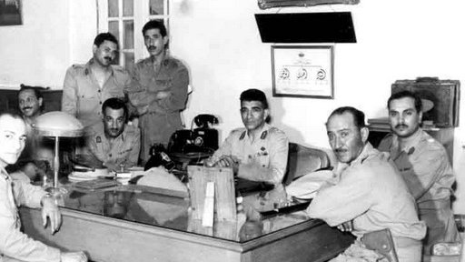 L'image montre un groupe de six hommes en uniforme militaire réunis autour d'une grande table en bois dans un bureau. Certains d'entre eux sont assis, tandis que d'autres se tiennent derrière. L'environnement décoré de manière classique inclut des éléments tels qu'une lampe de bureau et des tableaux accrochés au mur. L'atmosphère semble formelle, et les expressions des hommes varient entre concentration et sérieux.