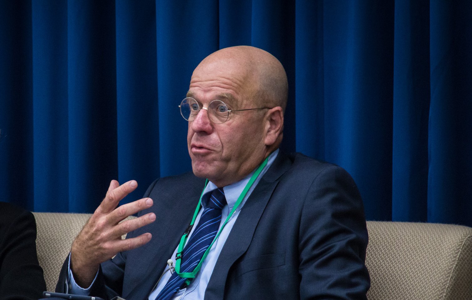 L'image montre un homme assis sur un canapé. Il a une tête chauve et porte des lunettes rondes. Il est vêtu d'un costume sombre avec une cravate. L'homme semble s'exprimer avec des gestes de la main, évoquant un discours ou une discussion. En arrière-plan, on peut voir un rideau bleu. L'atmosphère de l'image semble professionnelle.