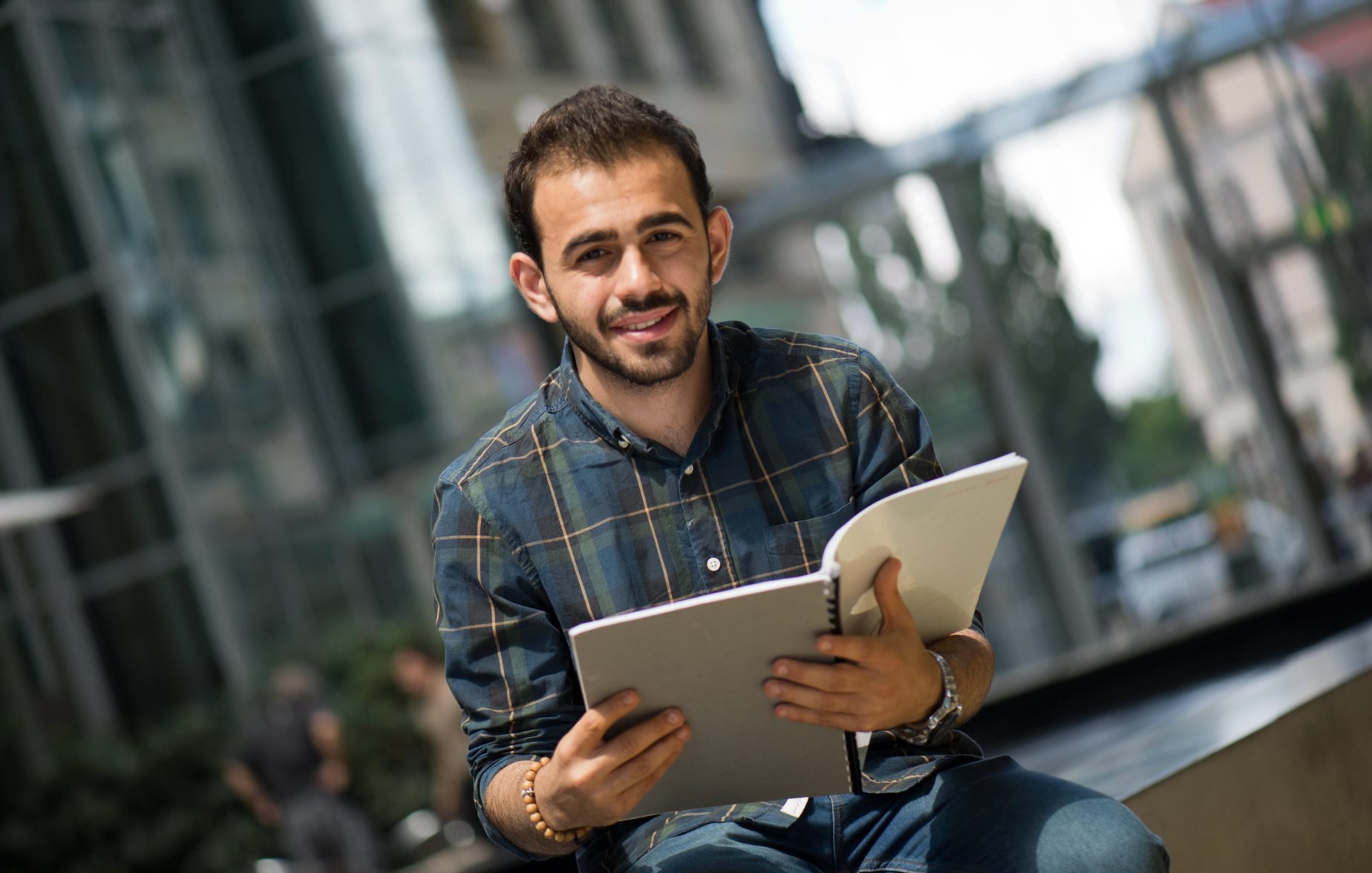 The image features a young man sitting outdoors, holding an open notebook or binder. He has a friendly smile and is wearing a plaid shirt. The background includes modern architecture and greenery, suggesting an urban setting. The atmosphere appears bright and inviting, indicating a pleasant day.