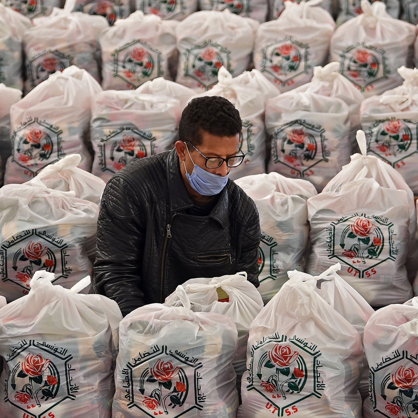 L'image montre un homme en train de travailler au milieu d'un grand espace rempli de sacs en plastique. Chaque sac est soigneusement empilé, arborant un motif de rose et des inscriptions en arabe. L'homme porte un masque et semble concentré sur sa tâche, probablement en train de trier ou de préparer quelque chose à l'intérieur des sacs. L'environnement est bien organisé, et la scène évoque un sentiment d'efficacité dans un contexte d'entraide ou de distribution.