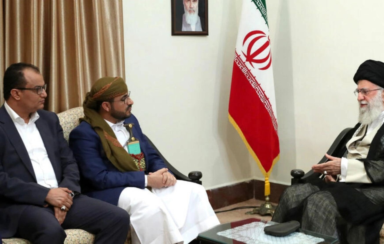 الصورة تُظهر ثلاثة رجال يجلسون في غرفة اجتماعات. في الخلفية، يوجد علم إيران وصورة كبيرة على الحائط. الرجل الموجود على اليمين يرتدي زيًا تقليديًا ويبدو أنه يتحدث مع الرجلين الآخرين، الذين يجلسون أمامه. يُظهر تعبيرهم اهتمامًا وتفاعلًا مع الحديث. الغرفة تبدو رسمية، مع أثاث بسيط وإضاءة دافئة.