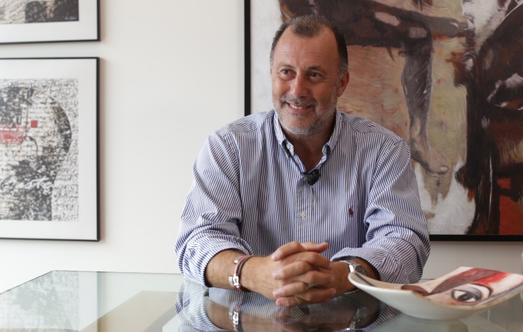 L'image montre un homme assis à une table en verre, semblant détendu et souriant. Il porte une chemise à rayures et des bracelets. En arrière-plan, on peut voir des œuvres d'art accrochées au mur, donnant une ambiance artistique à la pièce. Sur la table, il y a une assiette et des documents, ce qui suggère un contexte de discussion ou d'entretien. L'ensemble donne une impression de convivialité et d'élégance.