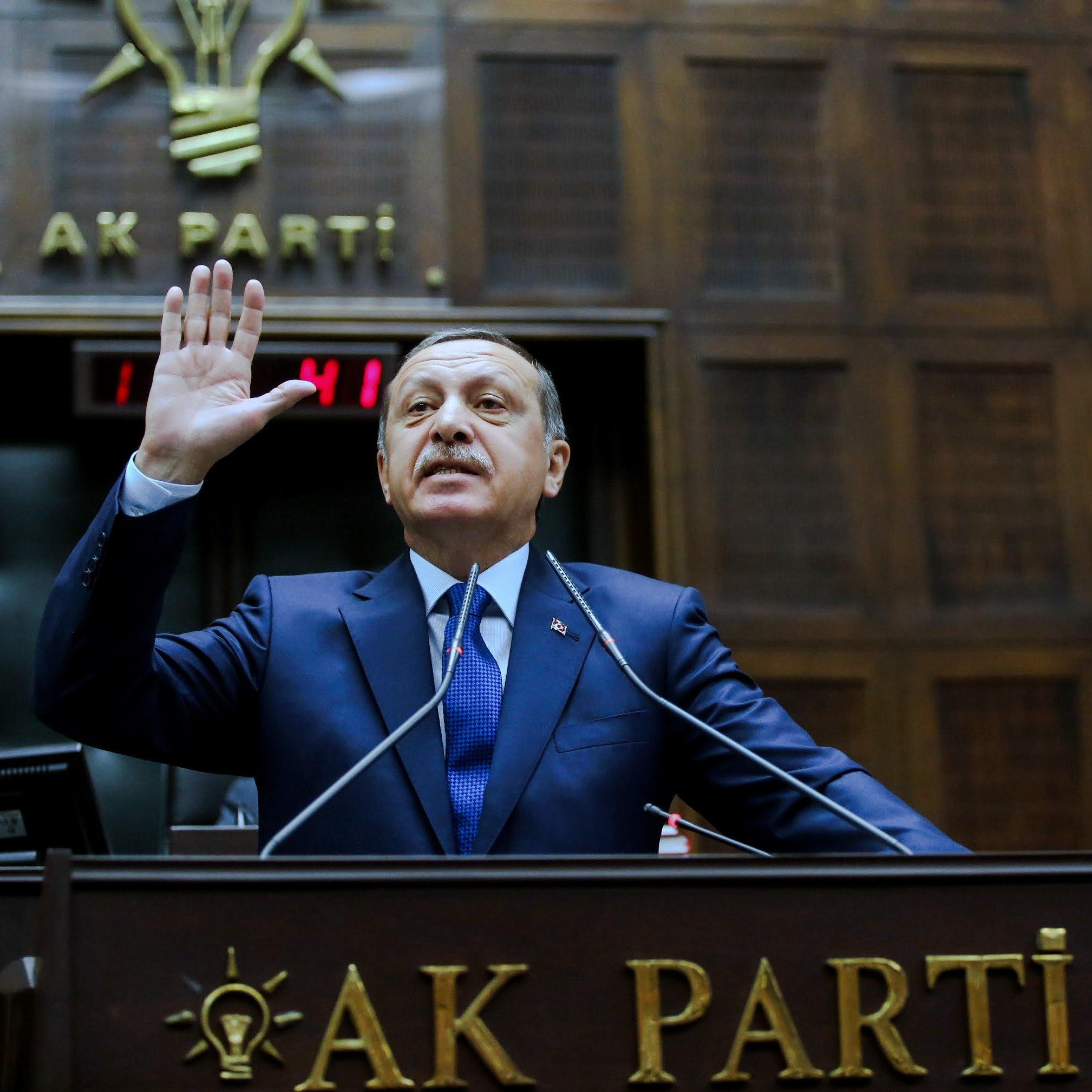 L'image montre un homme, probablement dans un contexte politique, levant la main en signe de serment ou de déclaration. Il est vêtu d'un costume sombre et d'une cravate. Derrière lui, on peut voir des éléments de décoration liés à un parti politique, avec un emblème et un drapeau. L'environnement semble être celui d'une assemblée ou d'un conseil, suggérant une atmosphère formelle et politique.