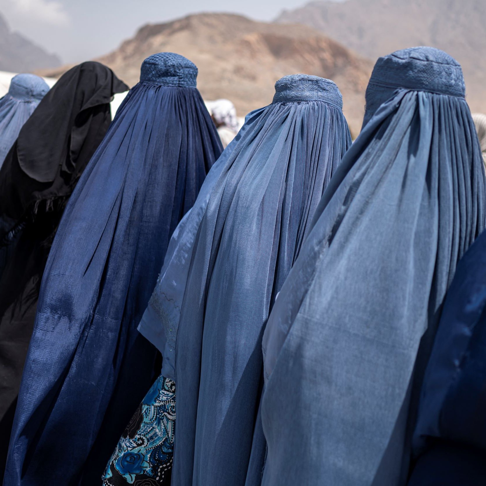 L'image montre un groupe de femmes de dos, vêtues de burqas de différentes nuances de bleu, se tenant en ligne. On peut également apercevoir une femme portant des vêtements noirs. En arrière-plan, on distingue des montagnes, et le ciel semble nuageux. L'ambiance générale semble être celle d'un rassemblement ou d'une rencontre.