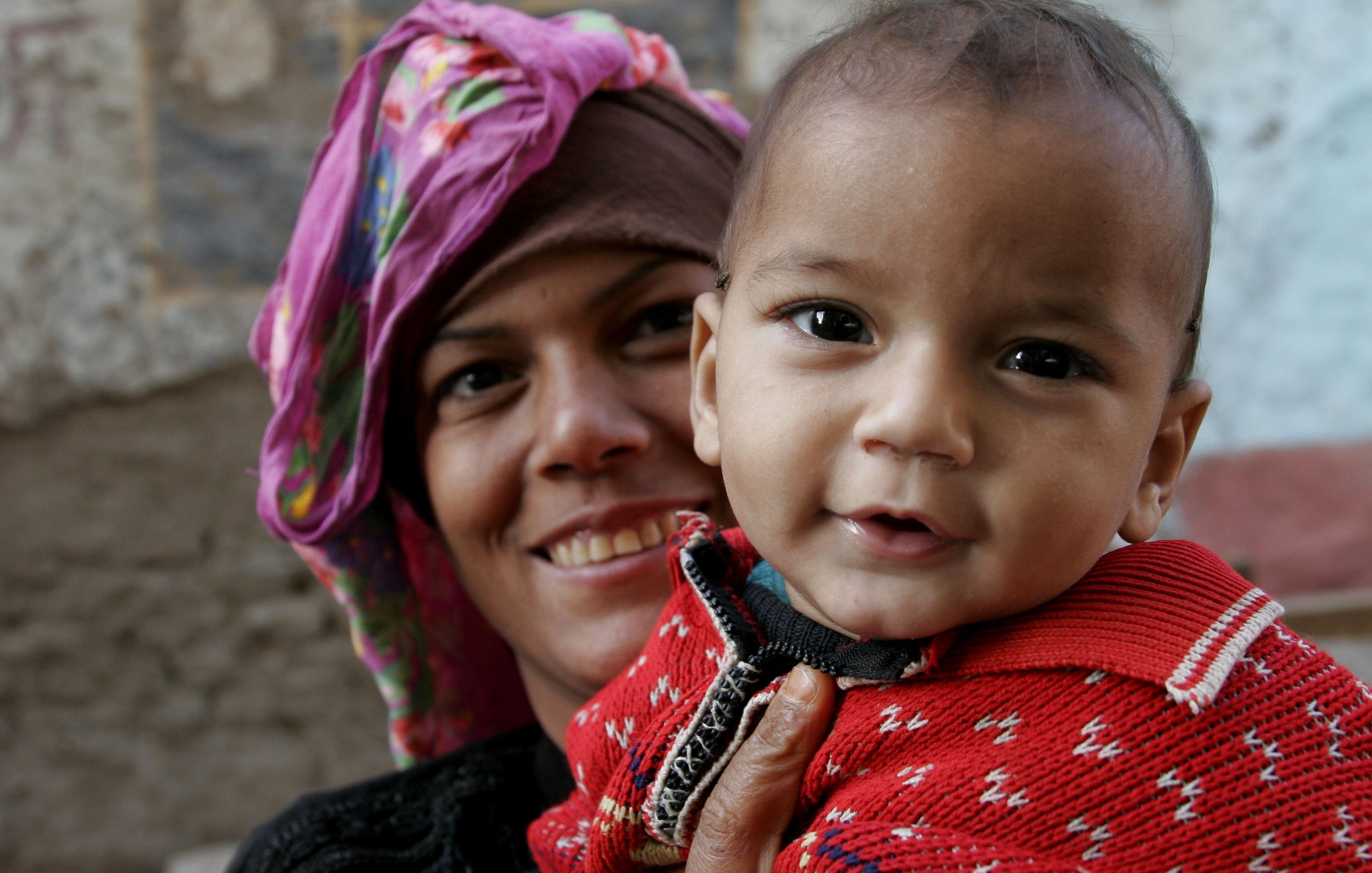 تظهر في الصورة امرأة تحمل طفلاً صغيراً. المرأة ترتدي حجاباً ملوناً وتبتسم، مما يعكس سعادة وتواصل مع الطفل. الطفل يرتدي سترة حمراء ذات نقش جميل ويبدو مبتسمًا أيضًا. خلفية الصورة تعكس بيئة بسيطة، مع جدران قديمة ذات ألوان طبيعية. الجو العام للصورة يوحي بالحب والحنان بين الأم وطفلها.