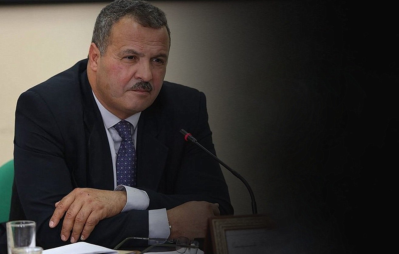 الصورة تظهر رجلًا يرتدي بدلة رسمية. يبدو أنه في اجتماع أو مناقشة، حيث يجلس أمام ميكروفون. يدي الرجل متقاطعتان أمامه، وهو يحدق في شيء ما بعناية. الخلفية غامقة قليلاً، مما يركز الانتباه على وجهه وتعبيراته.