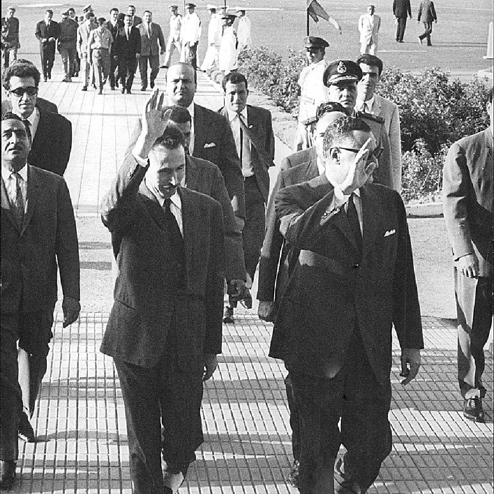 L'image montre un groupe de personnes marchant sur un chemin, avec plusieurs individus en costume. Deux hommes au premier plan saluent, vraisemblablement en se dirigeant vers une foule ou un événement. En arrière-plan, on peut apercevoir d'autres personnes et des éléments d'architecture, ainsi que des officiers en uniforme. L'atmosphère semble formelle et dynamique, indiquant une occasion spéciale.