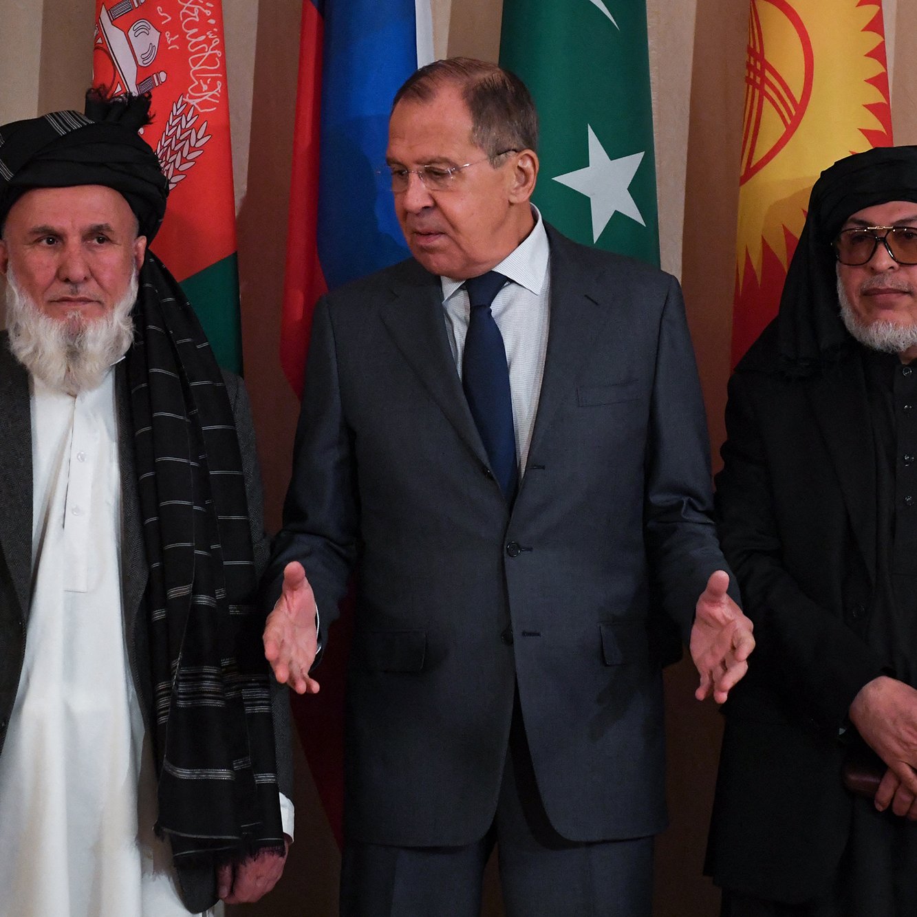 تظهر الصورة مجموعة من الرجال يقفون في مواجهة كاميرا. في الوسط، يوجد رجل يرتدي بدلة رسمية وهو يتحدث، بينما يقف على يساره رجل يرتدي زياً تقليدياً مع عمامة وشال، وعلى يمينه رجل آخر يرتدي ملابس مماثلة. خلفهم، تبرز مجموعة من الأعلام تمثل دول مختلفة، مما يوحي بأنهم في حدث رسمي أو مؤتمر دولي. تعكس تعابير الوجوه الوضع الجاد والتركيز على الحديث.