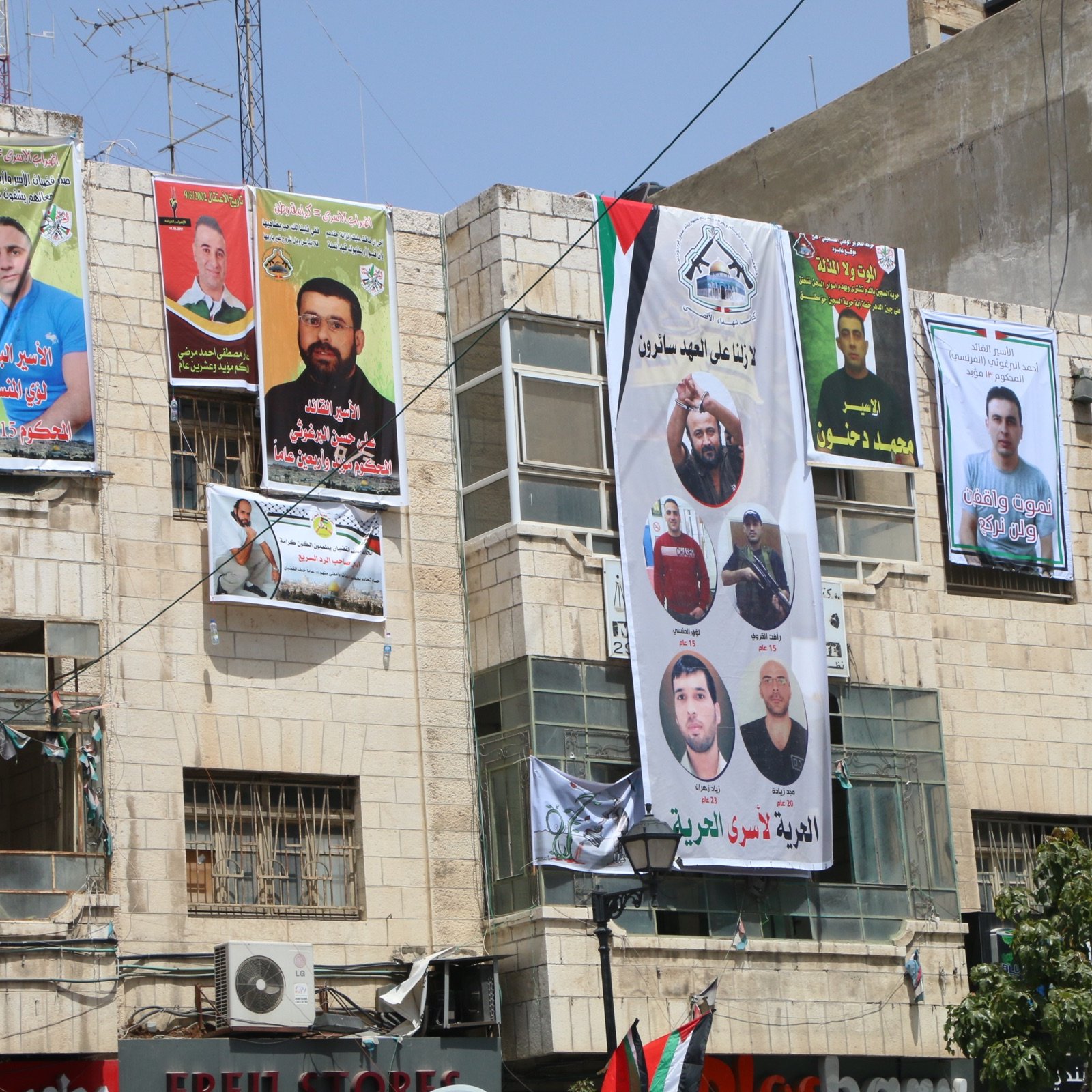 L'image montre un bâtiment avec plusieurs affiches et bannières accrochées sur sa façade. Ces affiches présentent des portraits de différentes personnes, probablement des figures politiques ou des militants, accompagnées de textes en arabe. Les couleurs des bannières sont vives, avec du jaune, du vert et du rouge, et beaucoup d'entre elles semblent commémorer ou soutenir des individus spécifiques. On peut également voir des drapeaux et d'autres éléments décoratifs liés à des mouvements politiques. L'environnement urbain donne une impression d'animation et de vie locale.