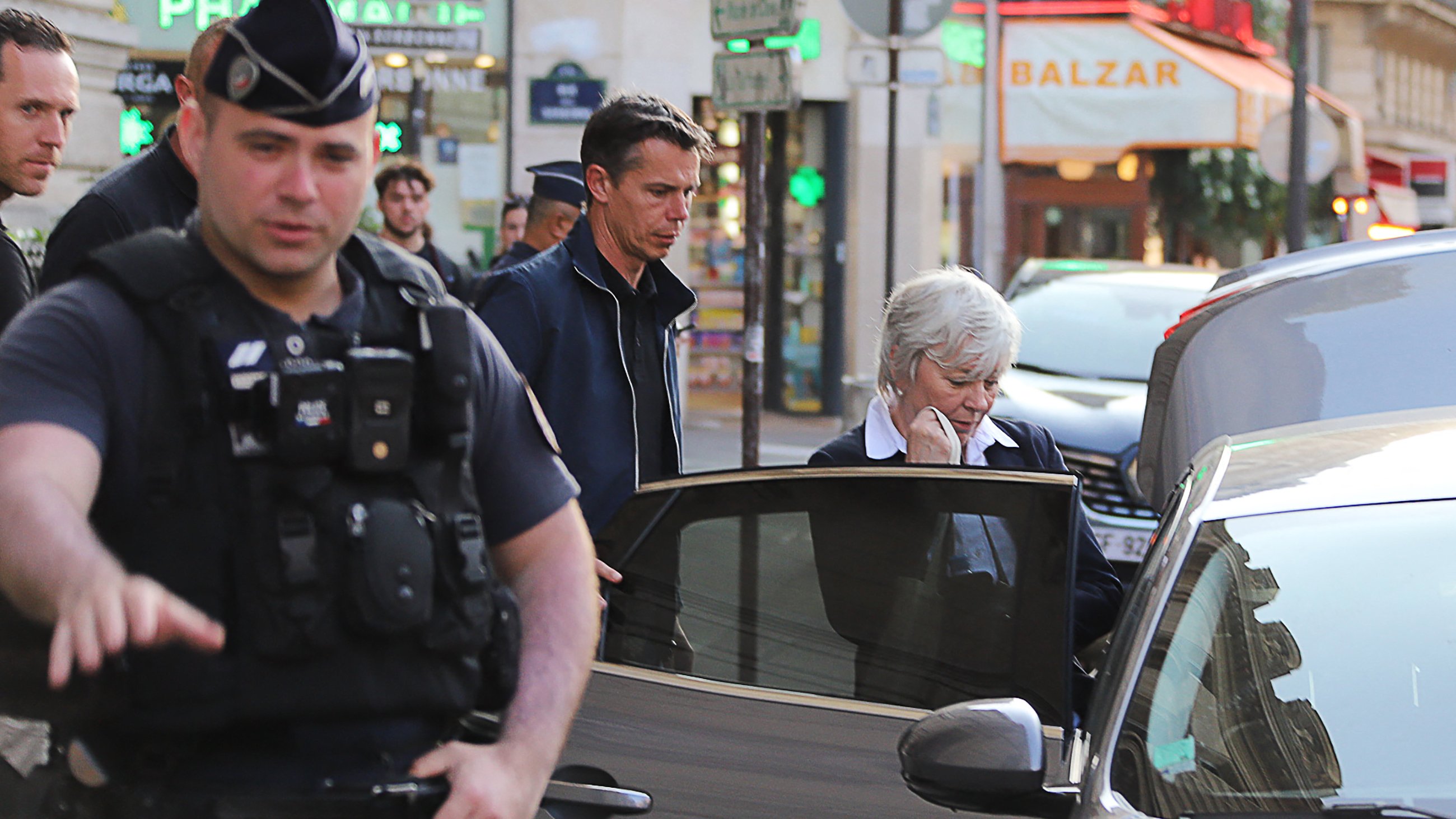 L'image montre une scène urbaine avec plusieurs personnes dans une rue. On peut voir un policier en uniforme à l'avant, surveillant la situation. À côté de lui, un homme et une femme semblent sortir d'une voiture. La femme est habillée de manière formelle et semble pensive. En arrière-plan, il y a plusieurs enseignes de magasins qui ajoutent à l'ambiance de la ville. Les lumières et l'architecture confèrent une atmosphère animée à la scène.