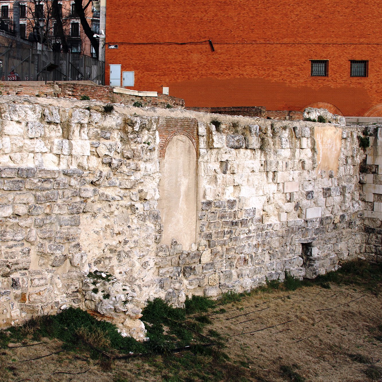 La imagen muestra una pared de piedra antigua, probablemente parte de una estructura histórica. La pared está compuesta de grandes bloques de piedra, algunos de los cuales están desgastados o dañados. En el centro, hay un espacio vacío en la pared que sugiere la existencia previa de una puerta o ventana. Al fondo, se puede ver un edificio de ladrillo rojo y un área de césped seco. Hay algunos restos de vegetación en la base de la pared, lo que añade un toque de naturaleza al entorno urbano.