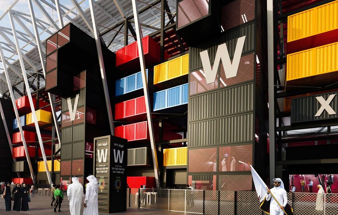 L'image montre une vue d'un bâtiment moderne, probablement un stade ou un centre événementiel, avec une architecture unique. Les murs extérieurs sont composés de containers colorés, alternant des teintes de jaune, rouge et bleu, donnant un aspect dynamique et contemporain. On distingue également des personnes en vêtements traditionnels se tenant près de l'entrée, ainsi que des éléments de sécurité. Le ciel est légèrement nuageux, ajoutant une ambiance particulière à la scène.