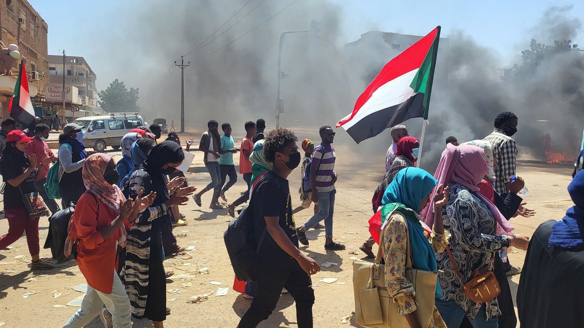Manifestation dans la rue, fumée, drapeau soudanais, personnes marchant ensemble.