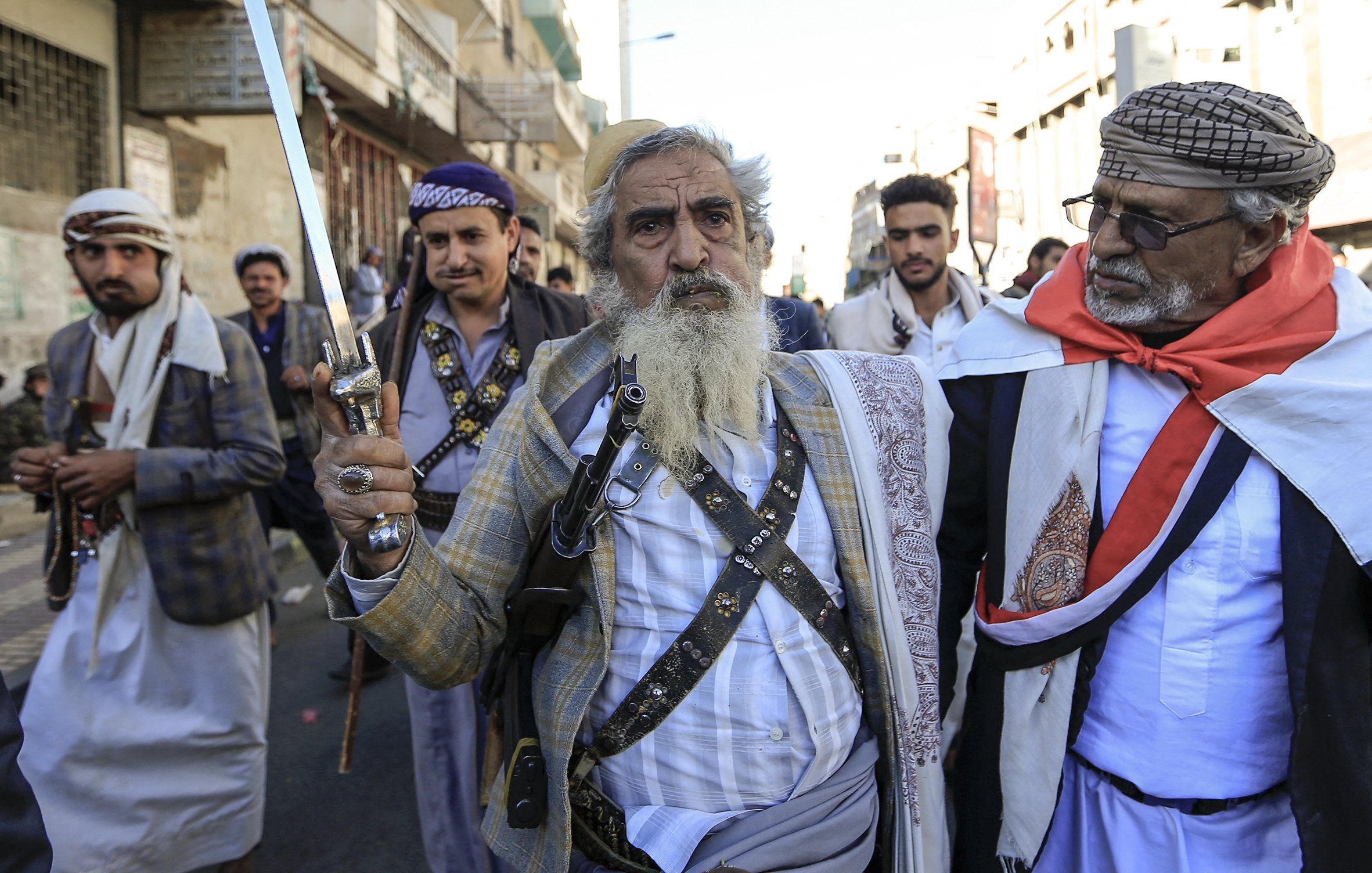 در این تصویر، چند مرد در حال راه رفتن در یک خیابان هستند. یکی از آن‌ها، که در پیش‌زمینه قرار دارد، دمی بلند و ریش سفیدی دارد و یک شمشیر در دست دارد. او لباس سنتی یمنی به تن کرده و به نظر می‌رسد که مقام یا اهمیت خاصی دارد. دیگر مردان نیز لباس‌های سنتی بر تن دارند و برخی از آن‌ها سلاح‌هایی در دست دارند. جو در تصویر به نظر مهیج و پر از نشاط است و نشان‌دهنده یک گردهمایی یا تجمع اجتماعی می‌باشد.