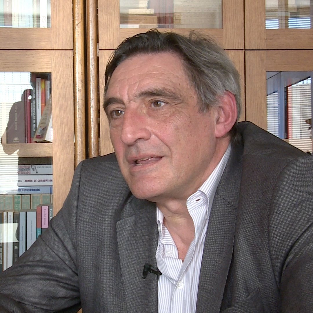 L'image montre un homme assis à une table, vêtu d'un costume gris. Il semble en train de parler ou de réfléchir, avec une expression sérieuse. En arrière-plan, on peut voir des étagères pleines de livres et des objets divers. La lumière naturelle entre par une fenêtre, créant un ambiance chaleureuse dans la pièce.