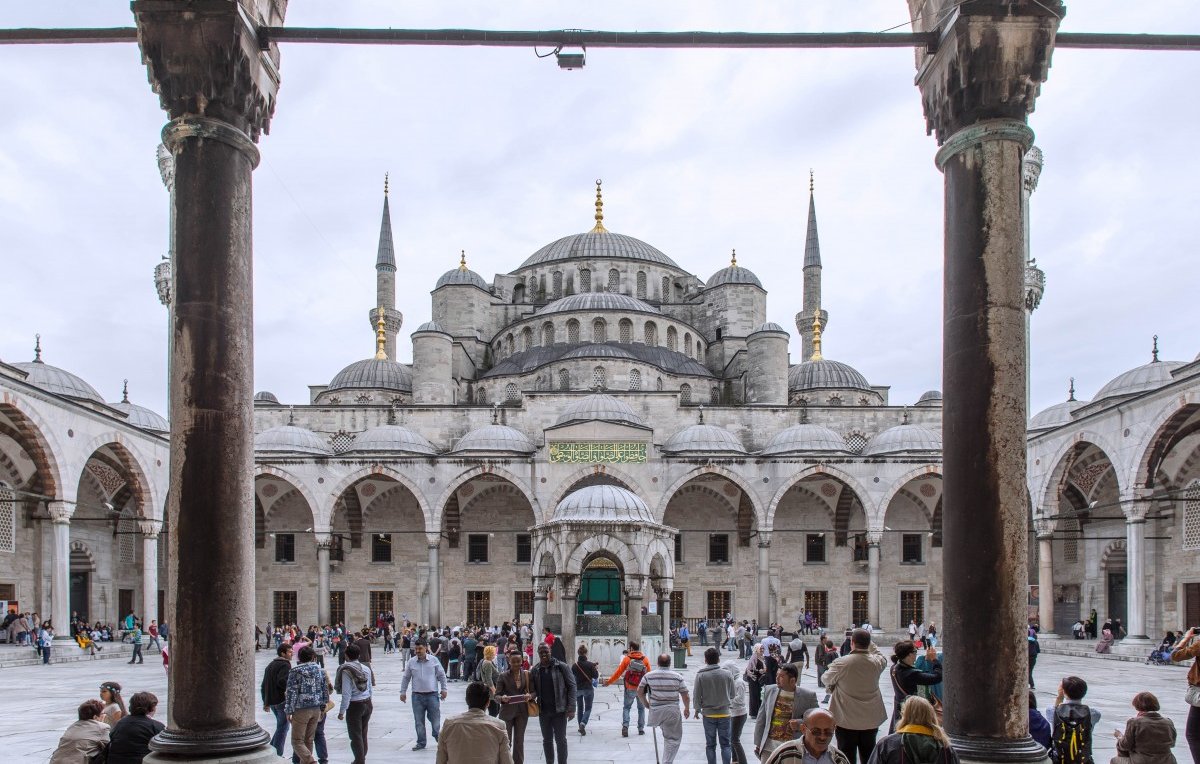 L'image montre une vue grandiose de la Mosquée bleue à Istanbul, encadrée par des arches majestueuses. Au premier plan, on peut voir un espace vaste et ouvert avec de nombreuses personnes qui se déplacent, créant une ambiance vivante. La mosquée elle-même se dresse en arrière-plan avec ses dômes et ses minarets élancés, sous un ciel nuageux qui ajoute une atmosphère particulière à la scène. Les détails architecturaux des colonnes et des arches sont également visibles, soulignant la beauté de cette œuvre historique.