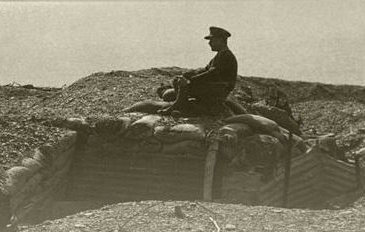 L'image représente un militaire assis sur un tas de terre ou de sacs de sable, surplombant une tranchée. Le soldat est vêtu d'un uniforme, et l'environnement semble être un champ de bataille ou une zone de guerre, probablement datant de la Première Guerre mondiale. Le ton de l'image est sépia, ce qui lui donne un aspect ancien et rétro, amplifiant le sentiment de nostalgie et de gravité de la situation.