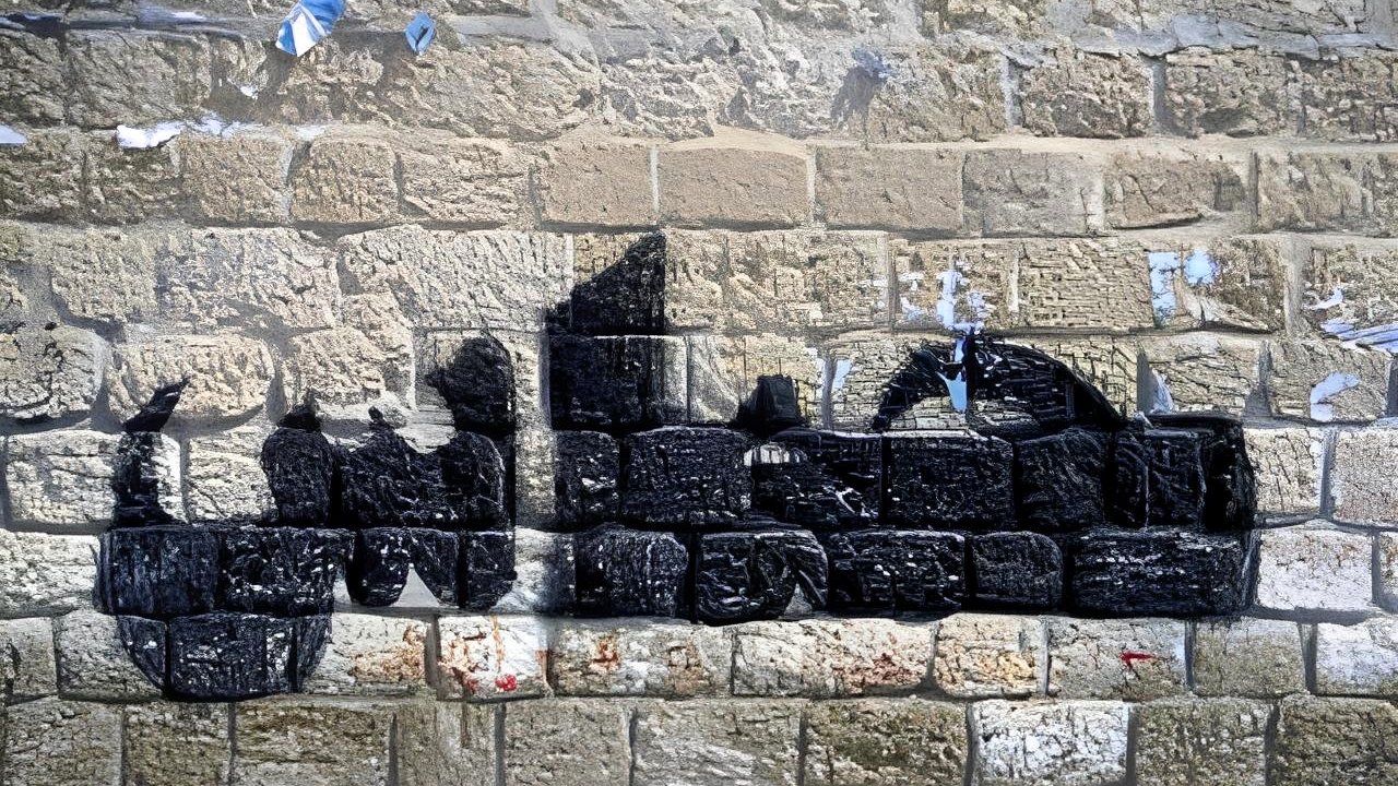 L'immagine mostra un murale su un muro di pietra. Il disegno rappresenta una silhouette di un veicolo, probabilmente un camion, realizzato con vernice nera. Le linee e le forme sono stilizzate, creando un contrasto con la texture della parete di pietra. Sul muro si notano anche resti di manifesti strappati e segni di usura, che aggiungono carattere e storia all'ambiente.