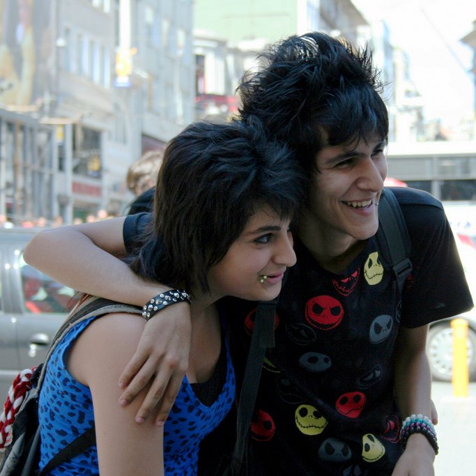 L'image montre deux jeunes personnes souriantes, posant côte à côte dans une rue animée. L'un des jeunes porte un t-shirt noir à motifs colorés, tandis que l'autre a un haut à motifs bleus. Ils semblent heureux et proches, et l'ambiance est vivante, avec des voitures et des piétons en arrière-plan. Les bâtiments en arrière-plan ajoutent à l'environnement urbain.