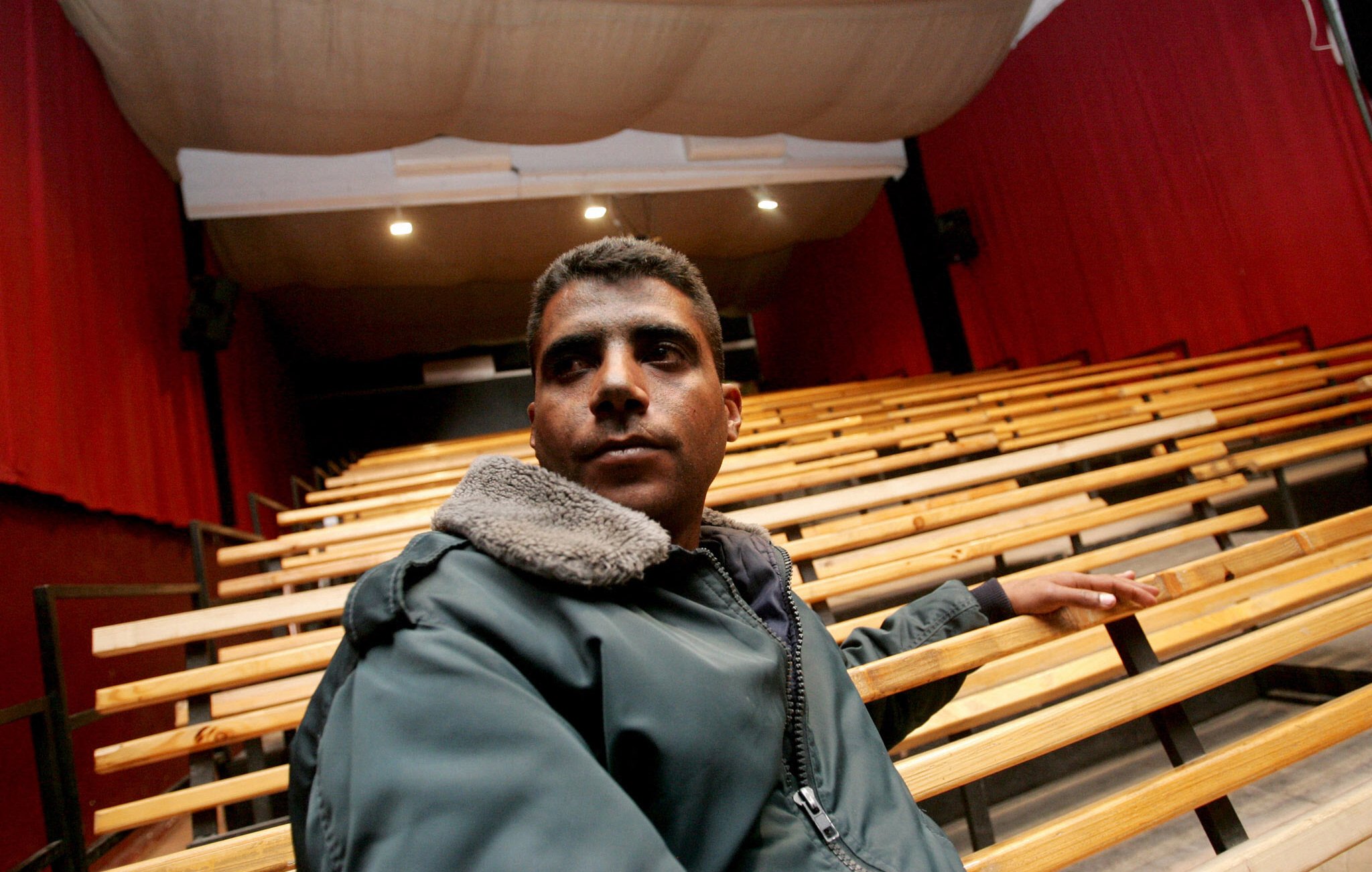 L'image montre un homme assis sur un banc dans un auditorium vide. L'homme porte une veste avec un col en fourrure et semble pensif, regardant vers l'avant. L'arrière-plan est composé de rangées de sièges en bois et de rideaux rouges, créant une atmosphère de théâtre ou de cinéma. La scène est bien éclairée, mettant en valeur l'homme et l'environnement autour de lui.