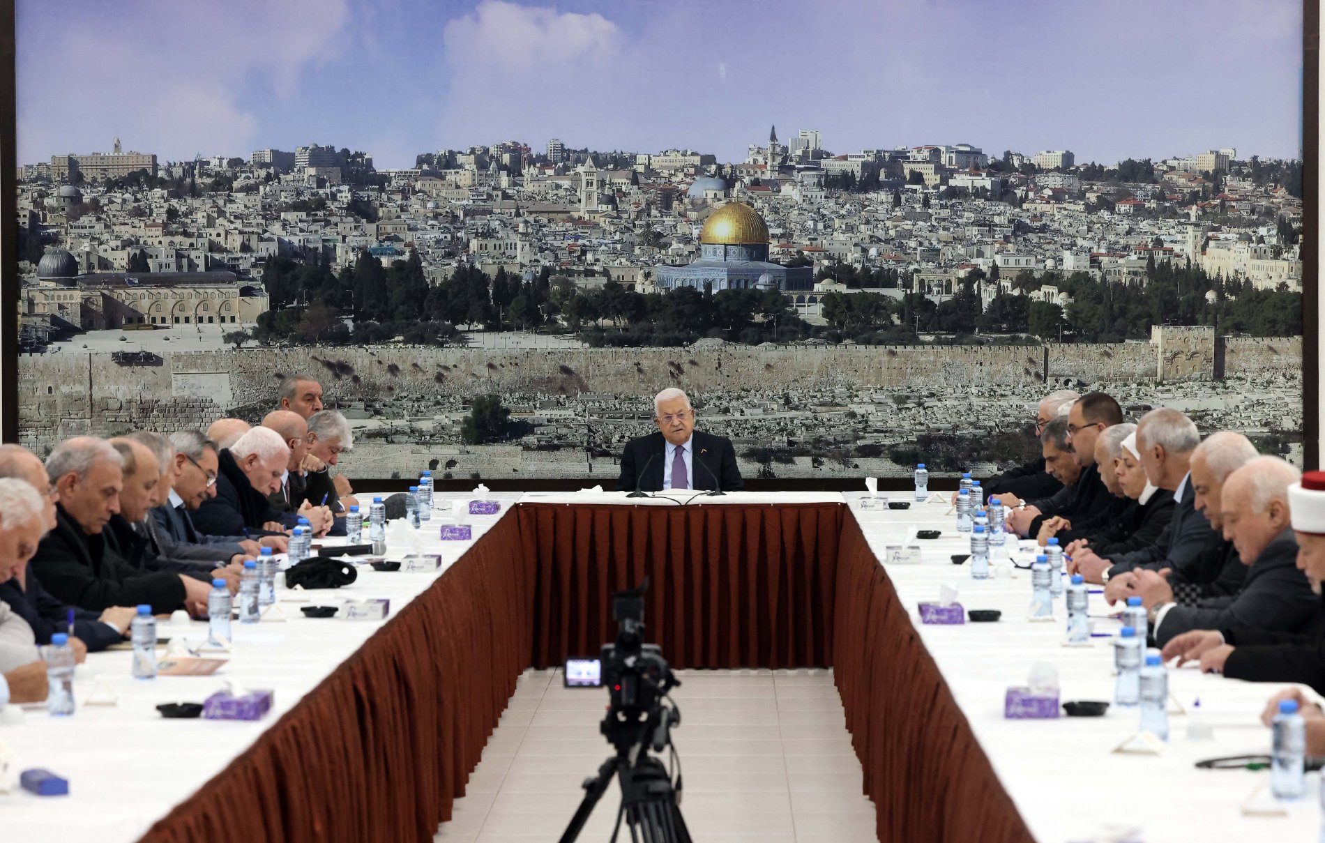 تظهر الصورة مجموعة من الأشخاص يجلسون حول طاولة كبيرة في قاعة اجتماع. في خلفية الصورة، يُمكن رؤية منظر لمدينة القدس مع قبة الصخرة. يبدو أن الجلسة تتعلق بقضية هامة، حيث يركز الجميع على شخص يجلس في المنتصف. الطاولة مرتبة بشكل مرتب، مع وجود زجاجات مياه وأكواب على الطاولة. يتمحور اللقاء حول موضوع قد يكون له تأثير كبير على المجتمع أو السياسة.