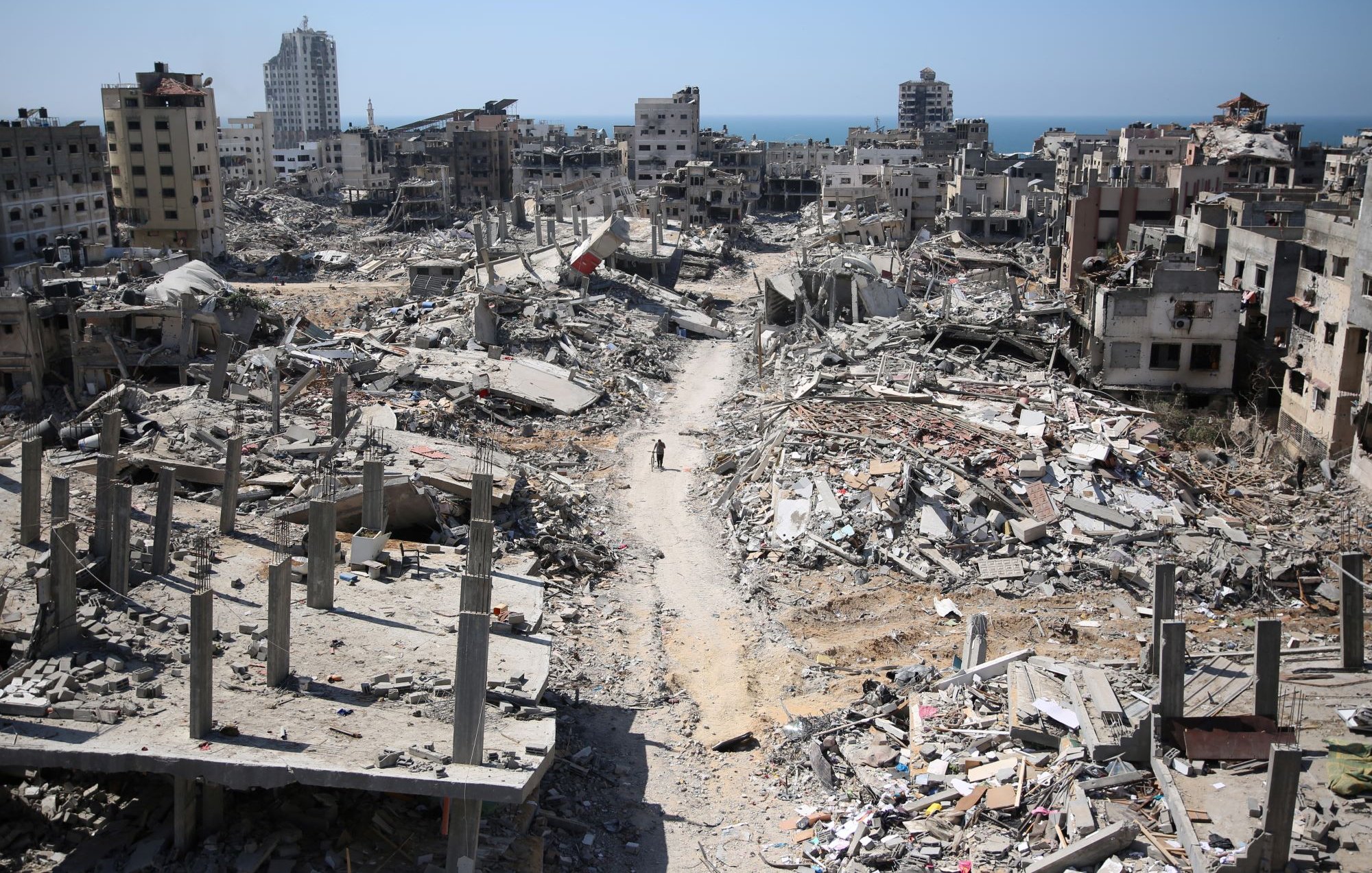 L'immagine mostra un paesaggio di devastazione, con edifici in rovina e macerie sparse in tutta l'area. Le strutture, una volta abitate, sono ridotte a scheletri di cemento e detriti. In lontananza, si scorge il mare, che contrasta con la desolazione del terreno. Una persona si muove tra le macerie, evidenziando la vastità della distruzione. Il cielo è sereno, creando un contrasto inquietante con la scena di desolazione sottostante.