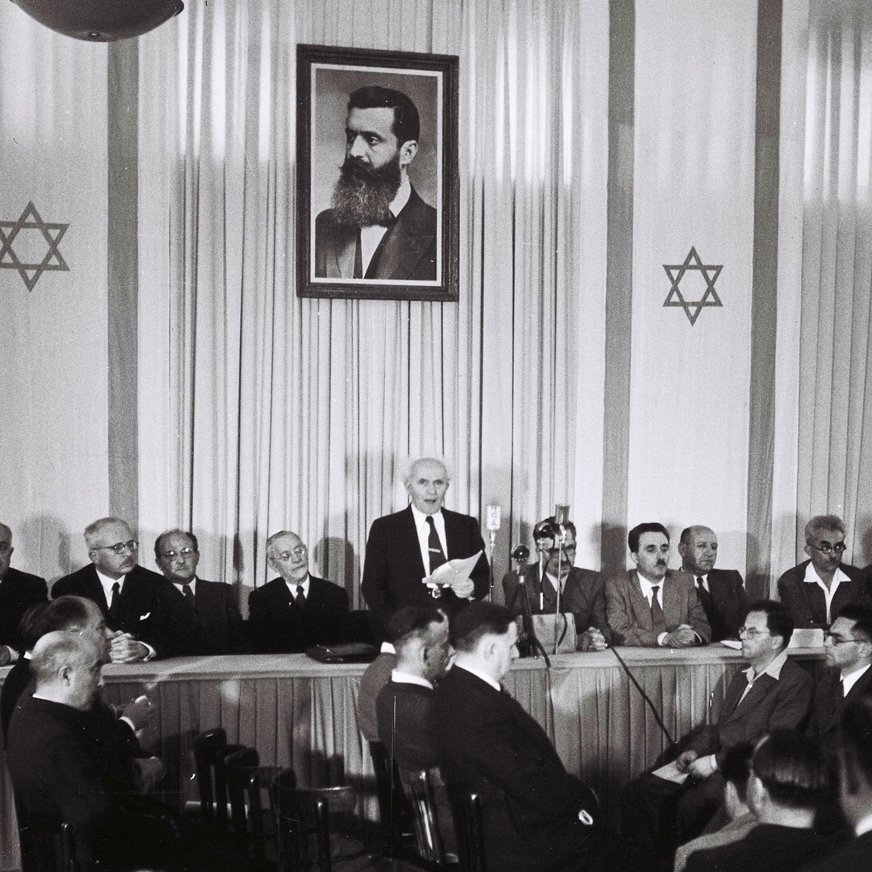 L'image montre une scène à l'intérieur d'une salle de réunion. Au fond, il y a un grand portrait en noir et blanc d'un homme, probablement une figure historique importante, entouré d'étoiles de David. Sur la scène, plusieurs hommes sont assis, en costume, tandis qu'un orateur se tient debout, s'adressant à l'assemblée. L'ambiance semble formelle, indiquant un événement significatif. Les participants, qui sont assis face à la scène, apparaissent attentifs à la présentation en cours.